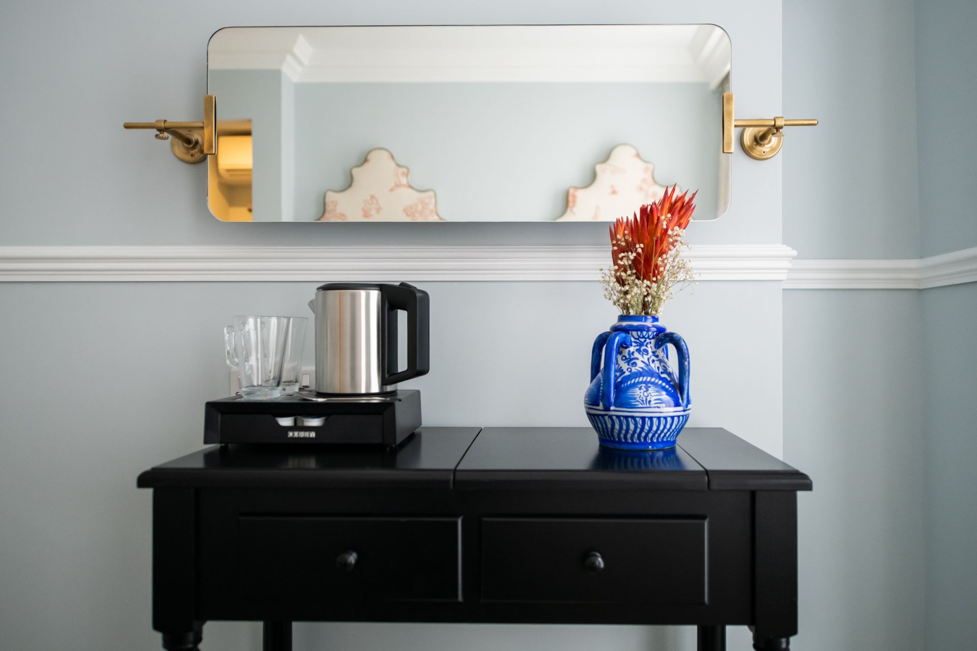 Black table with kettle, glasses, blue vase with flowers, under a gold-framed mirror on a blue wall.