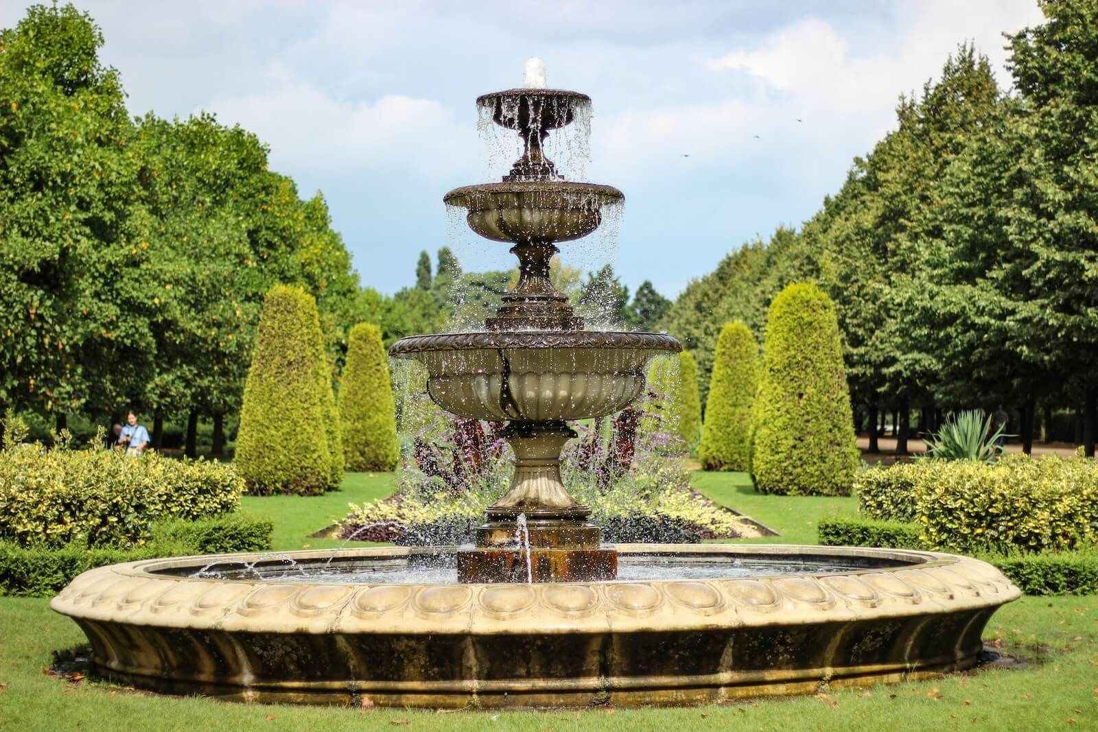 Ornate tiered fountain in a manicured garden, water spraying, surrounded by green shrubs and trees.