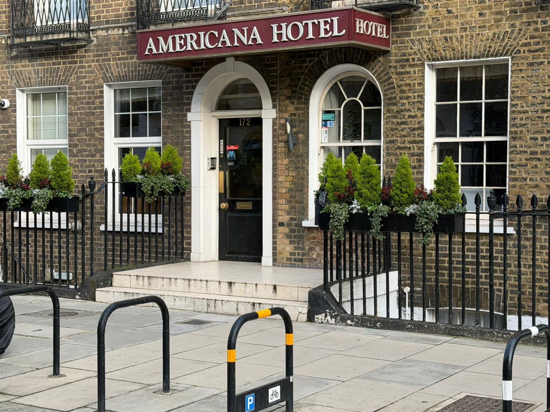 Americana Hotel exterior, brick building, arched doorway, steps, black wrought iron fence, small green trees in planters.