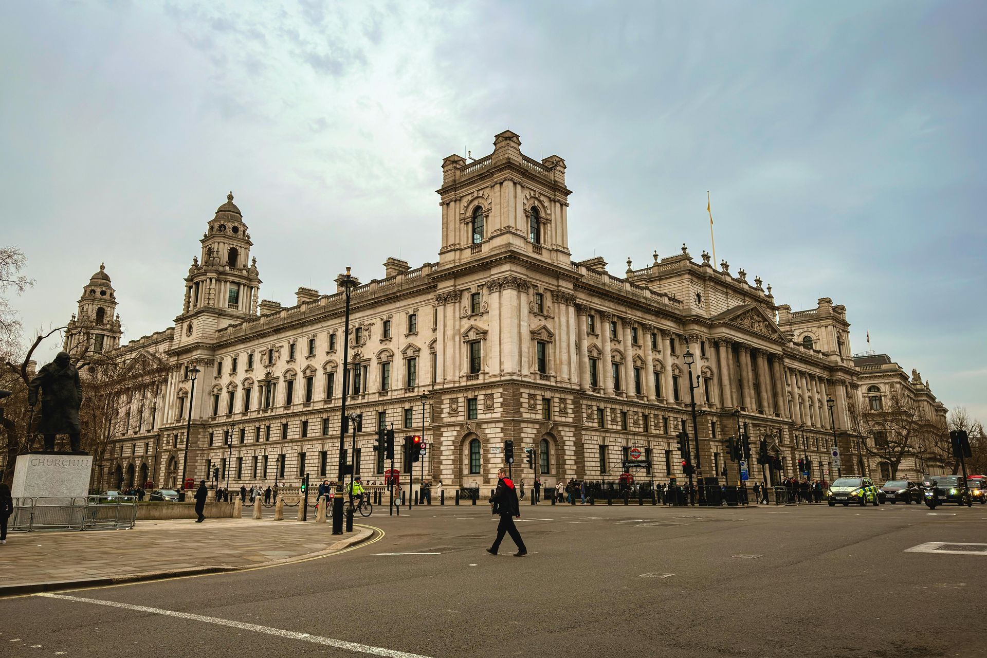 Exterior view of a large, ornate building with architectural details. A person crosses the street. Overcast sky.