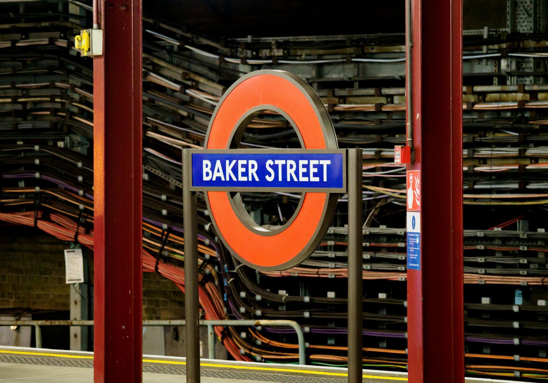 Baker Street London Underground station sign. Orange circle with blue text, red support poles, and cable backdrop.