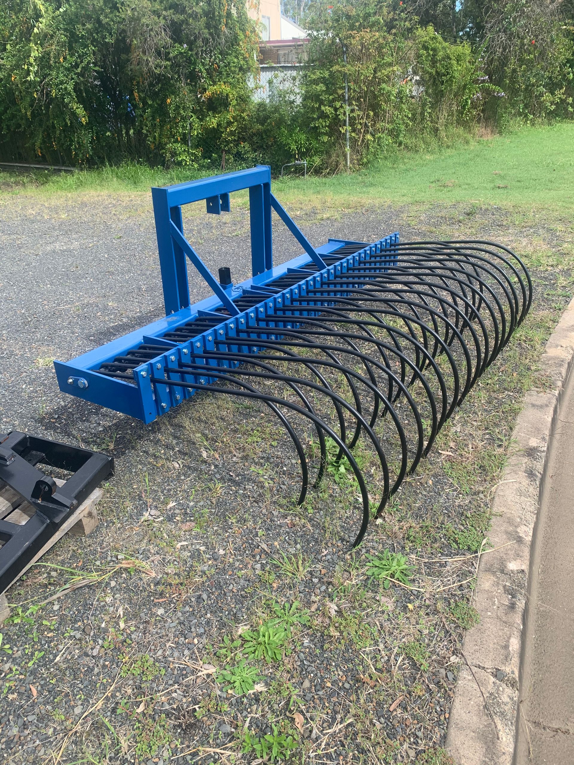 A Blue Rake is Sitting on the Ground Next to a Concrete Curb — Wingham Farm Machinery in Wingham, NSW