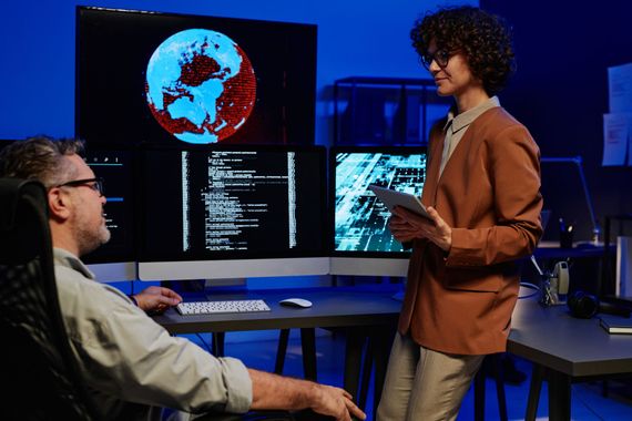 a man is sitting in front of a computer while a woman stands next to him holding a tablet | Apple Security Solutions