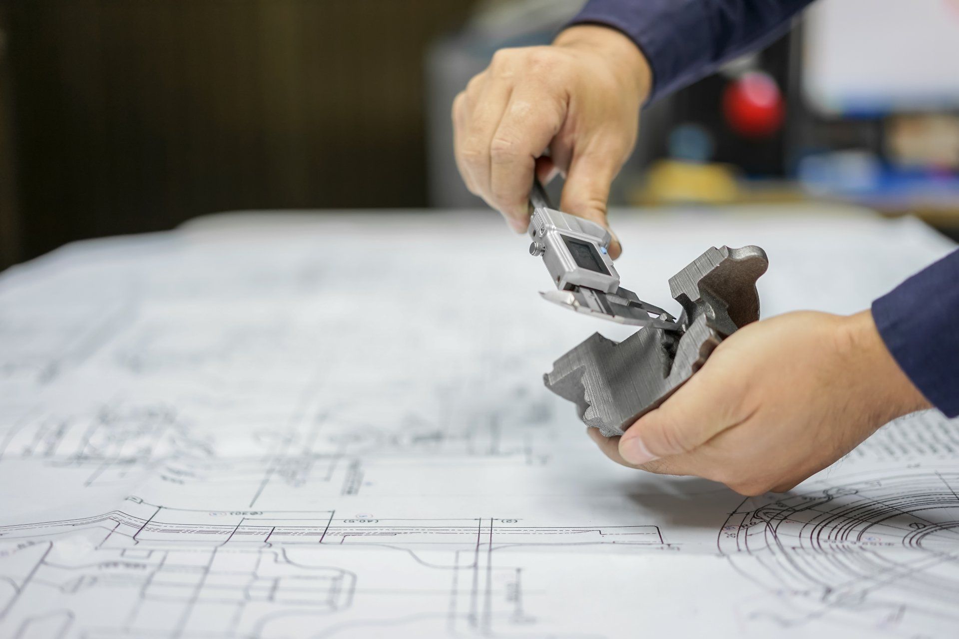 Eine Person hält vor einer Blaupause ein Stück Metall in den Händen.