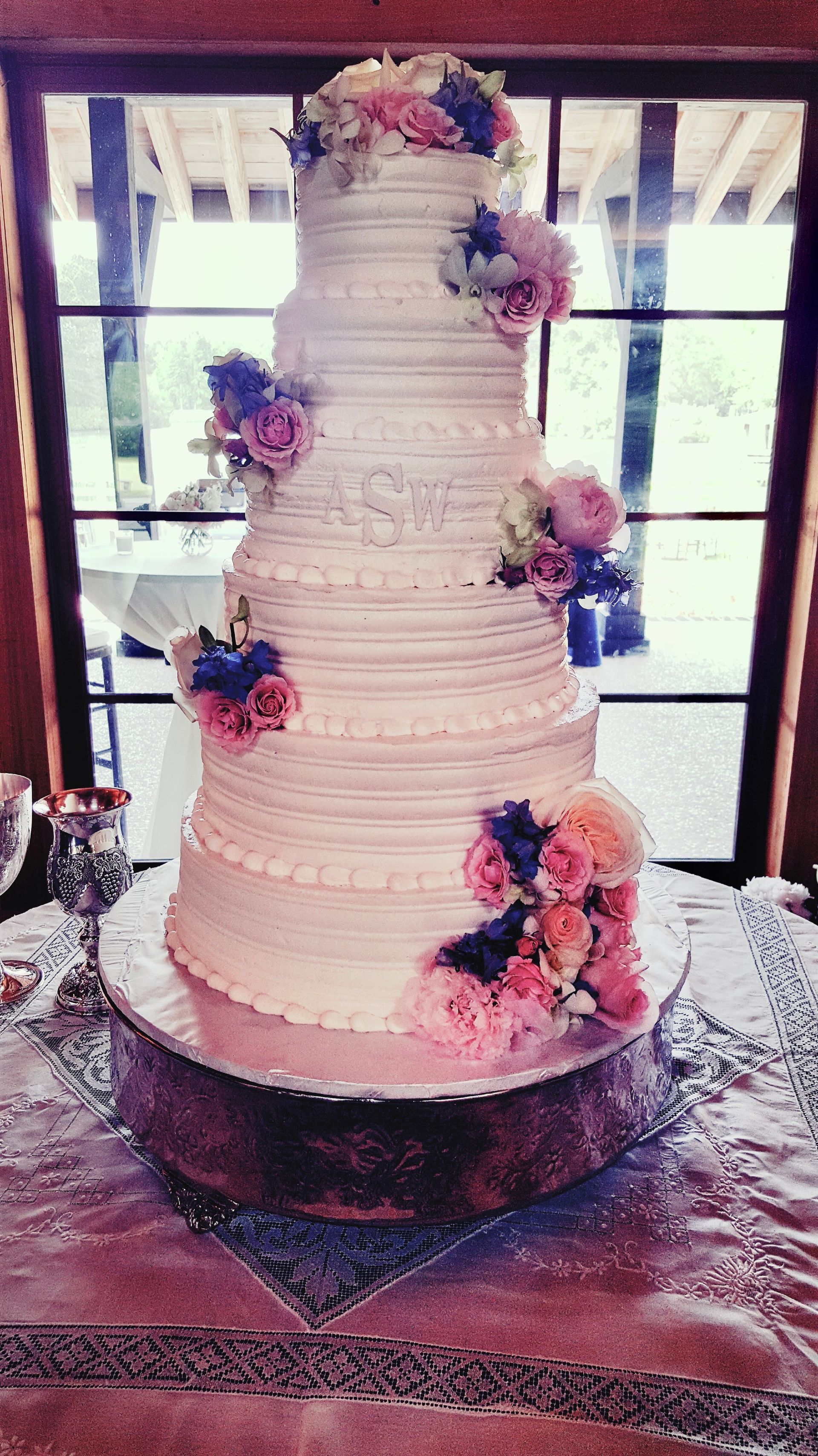 A tall, tiered wedding cake decorated with pink and blue flowers on a wooden cake stand in front of a window.