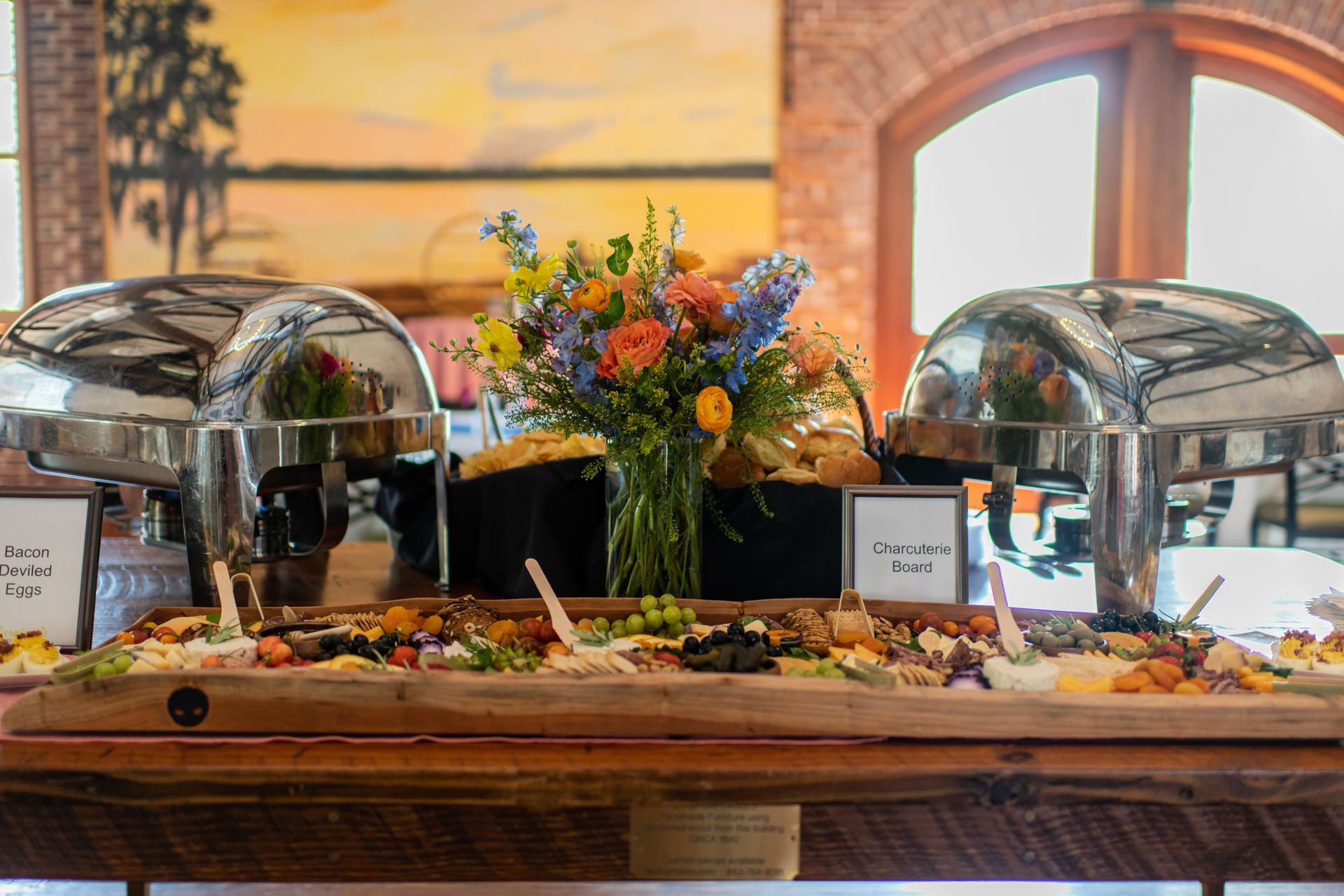 A buffet setup with a charcuterie board and covered food warmers, a floral arrangement in the center, set in a room with large windows and art.