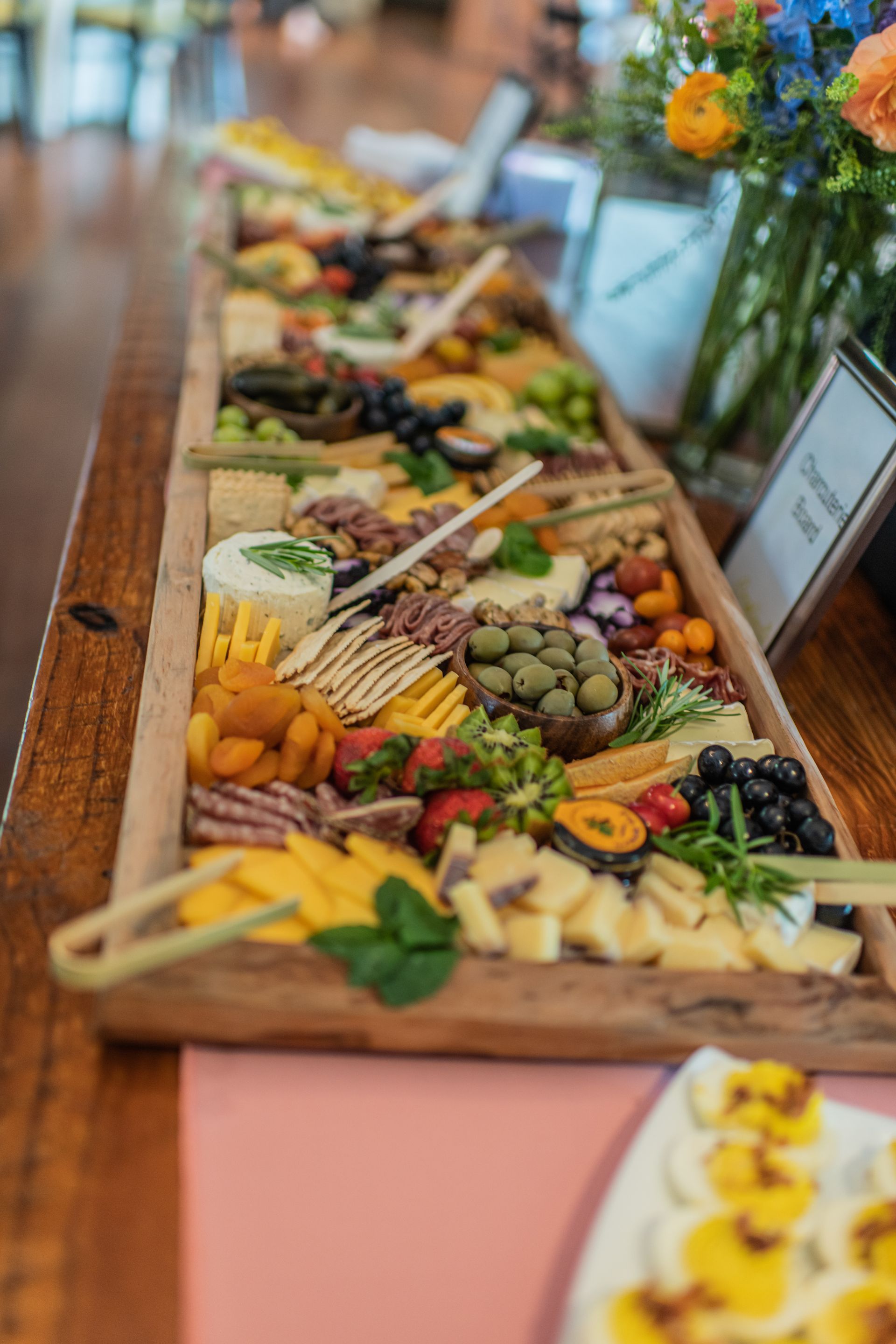 A long wooden charcuterie board displays various cheeses, fruits, and meats. The spread is set on a pink tablecloth.