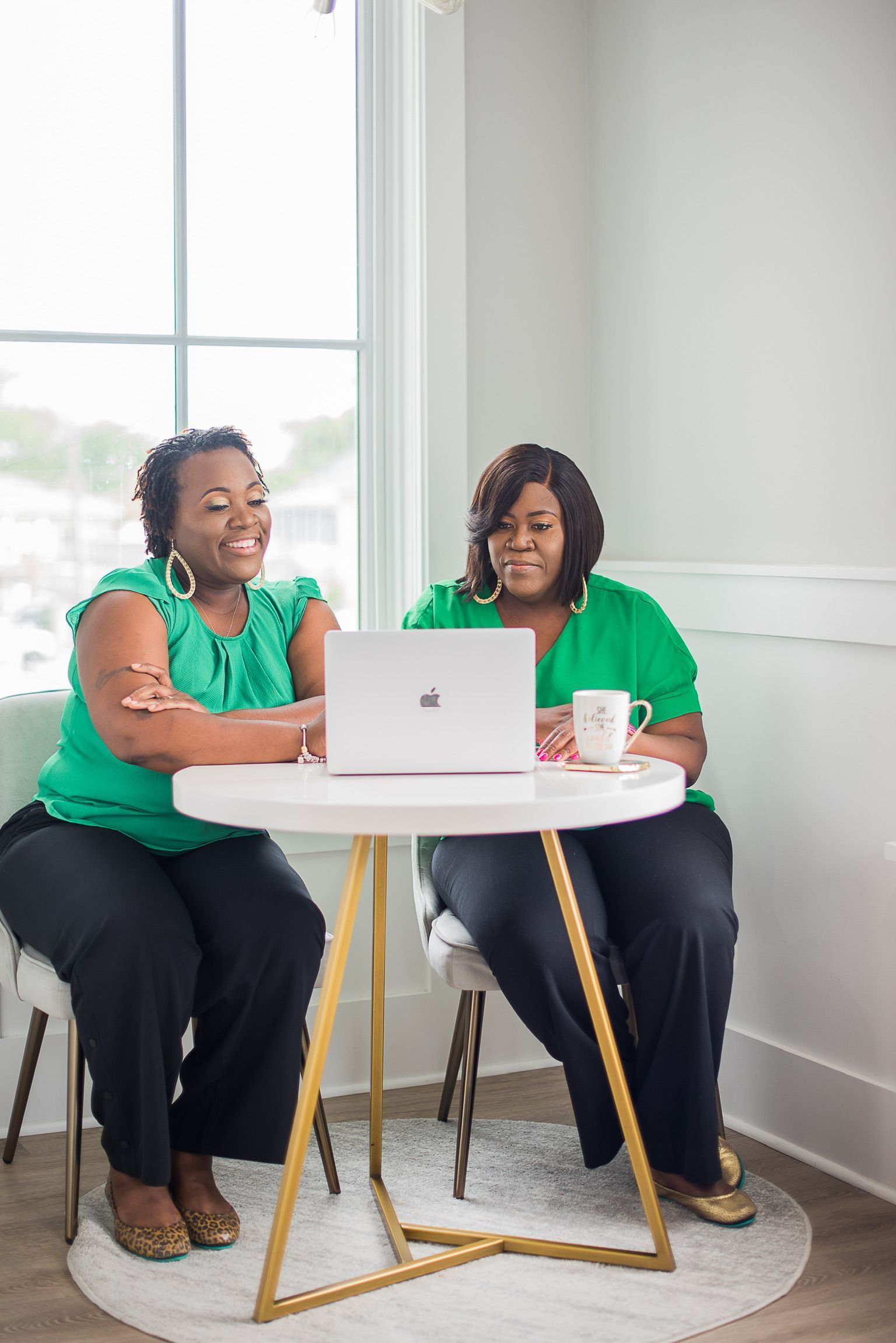 Two women in green shirts and black pants sit at a small round table with a laptop and coffee cup, smiling. They're near a window.