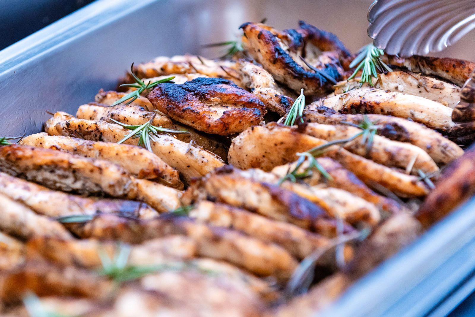 Grilled chicken strips in a metal serving dish, garnished with sprigs of rosemary.
