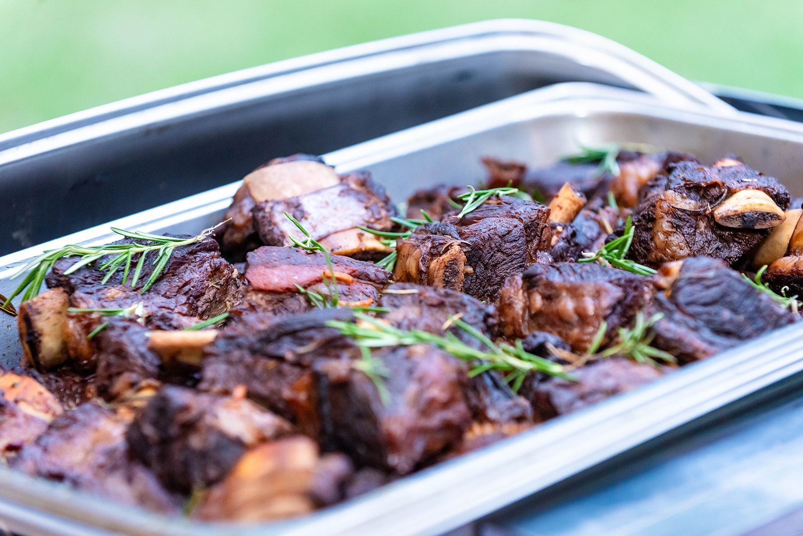 A metal serving tray filled with braised short ribs, garnished with fresh rosemary.