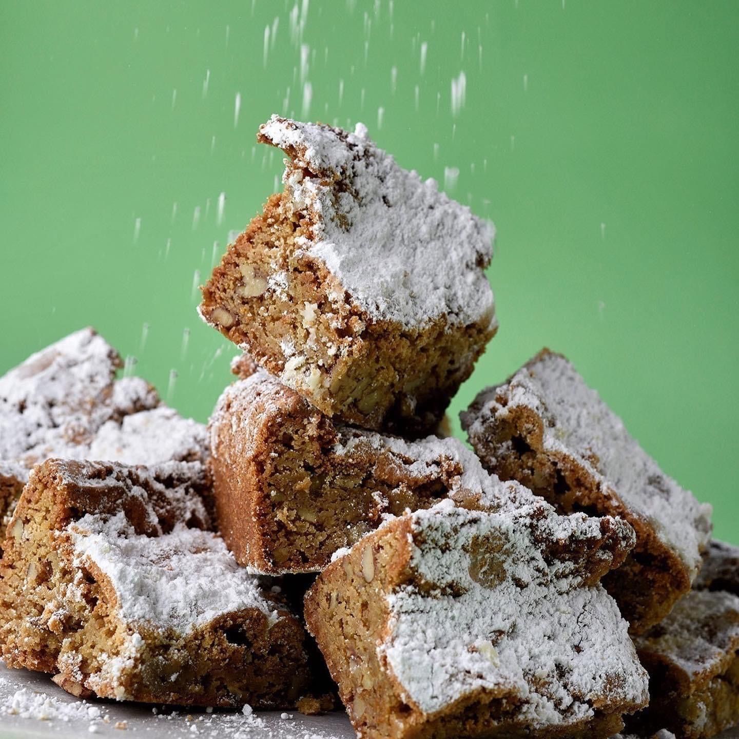 Pile of square baked goods dusted with powdered sugar, with more sugar falling from above against a green backdrop.
