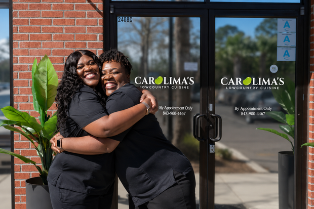 Two people hugging outside a brick storefront with glass doors and yellow lettering
