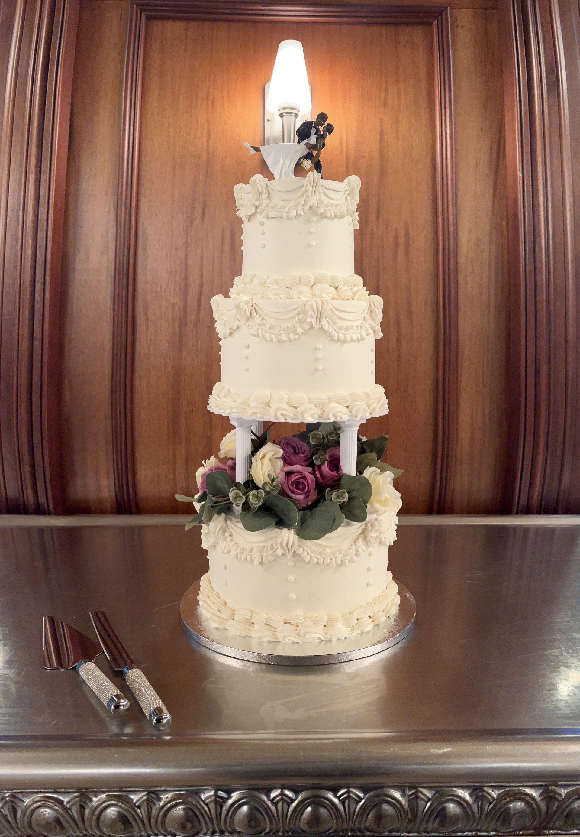 Four-tiered wedding cake with floral and white icing details. Cake knife set lays in front, with a wooden background.