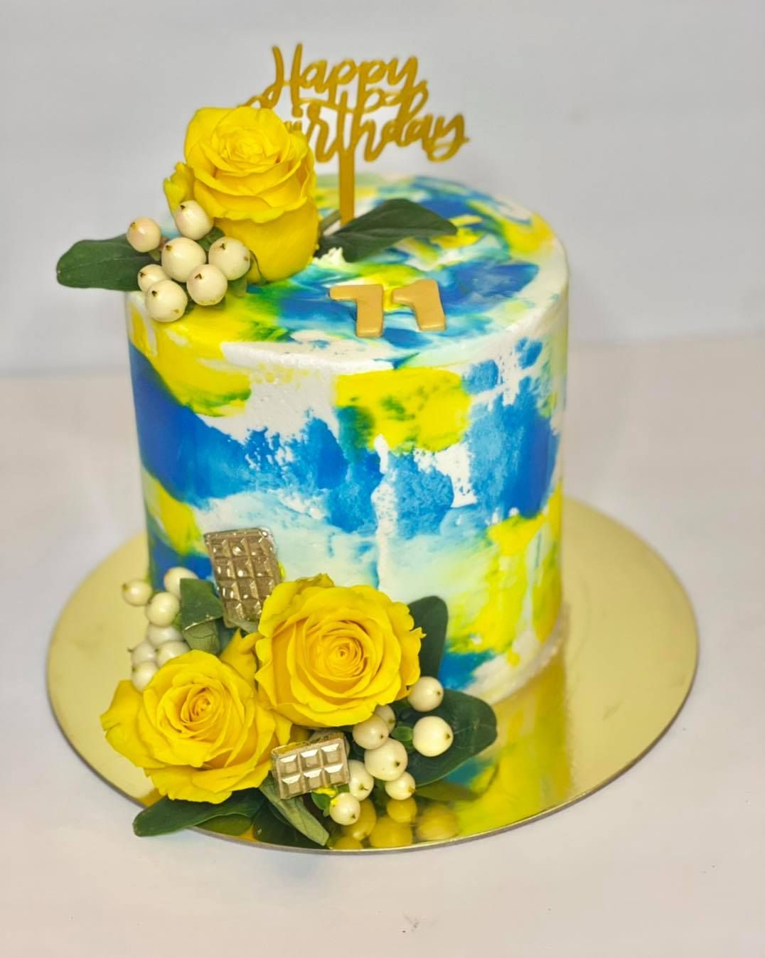 A celebratory cake with yellow and blue frosting, decorated with yellow roses, white berries, and a 