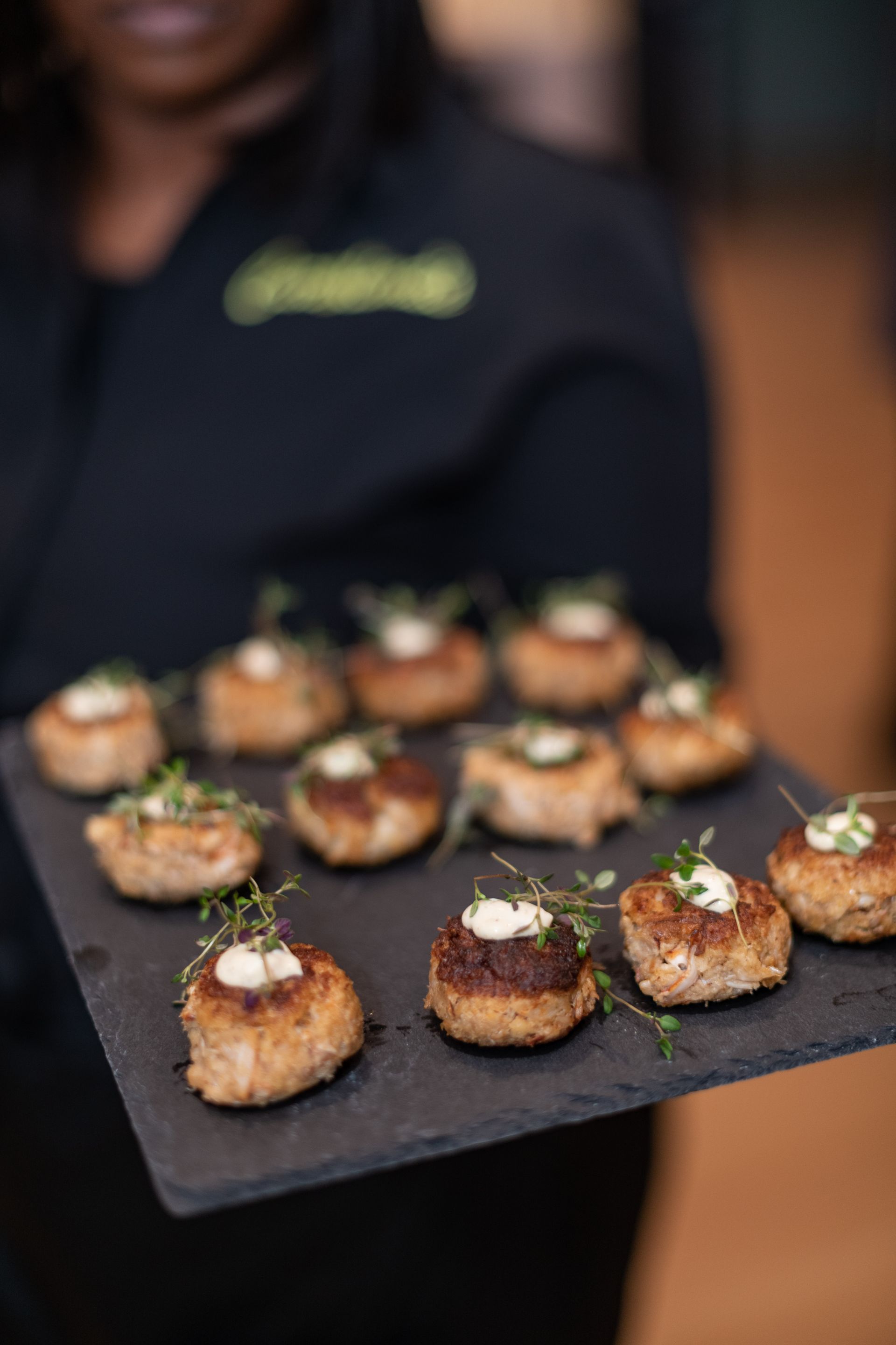 Server holding a platter of appetizers: round, browned food with a dollop of white sauce and sprigs of green herbs on top.