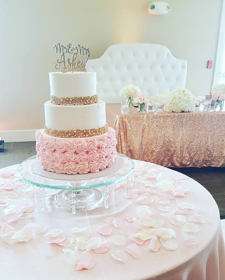 Three-tiered wedding cake with pink rose petals and gold accents, on a table decorated with rose petals.