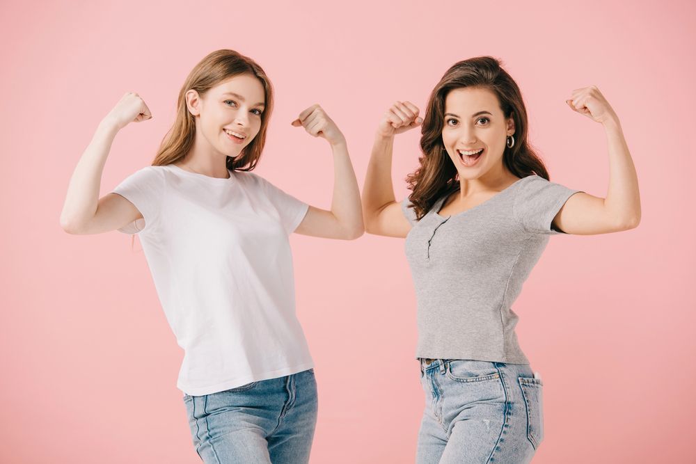Dos mujeres flexionan los brazos, sonriendo, sobre un fondo rosa.