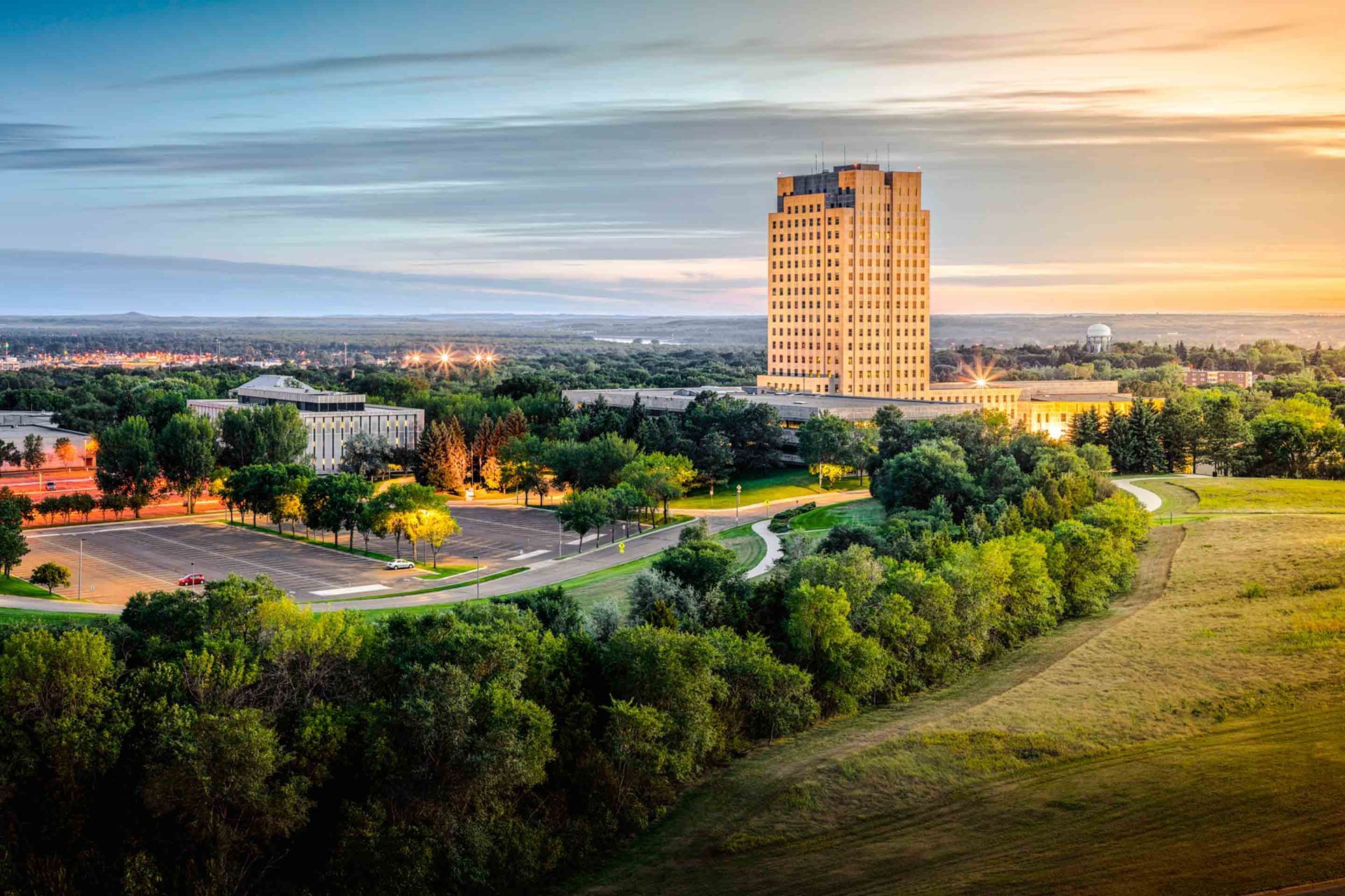 Tall building dominates a campus at sunset, surrounded by trees and a grassy field.