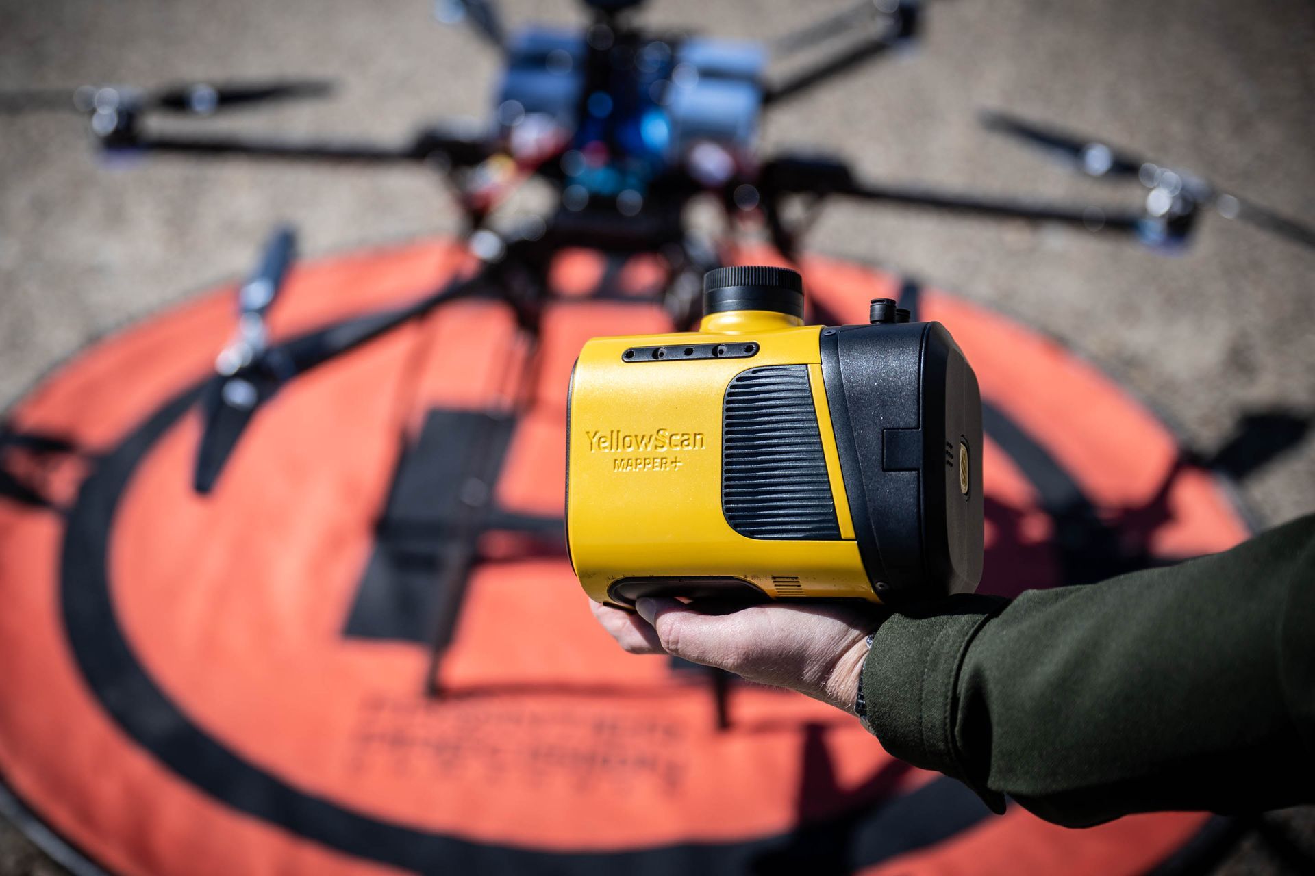 Person holding a yellow device in front of a drone on an orange landing pad.