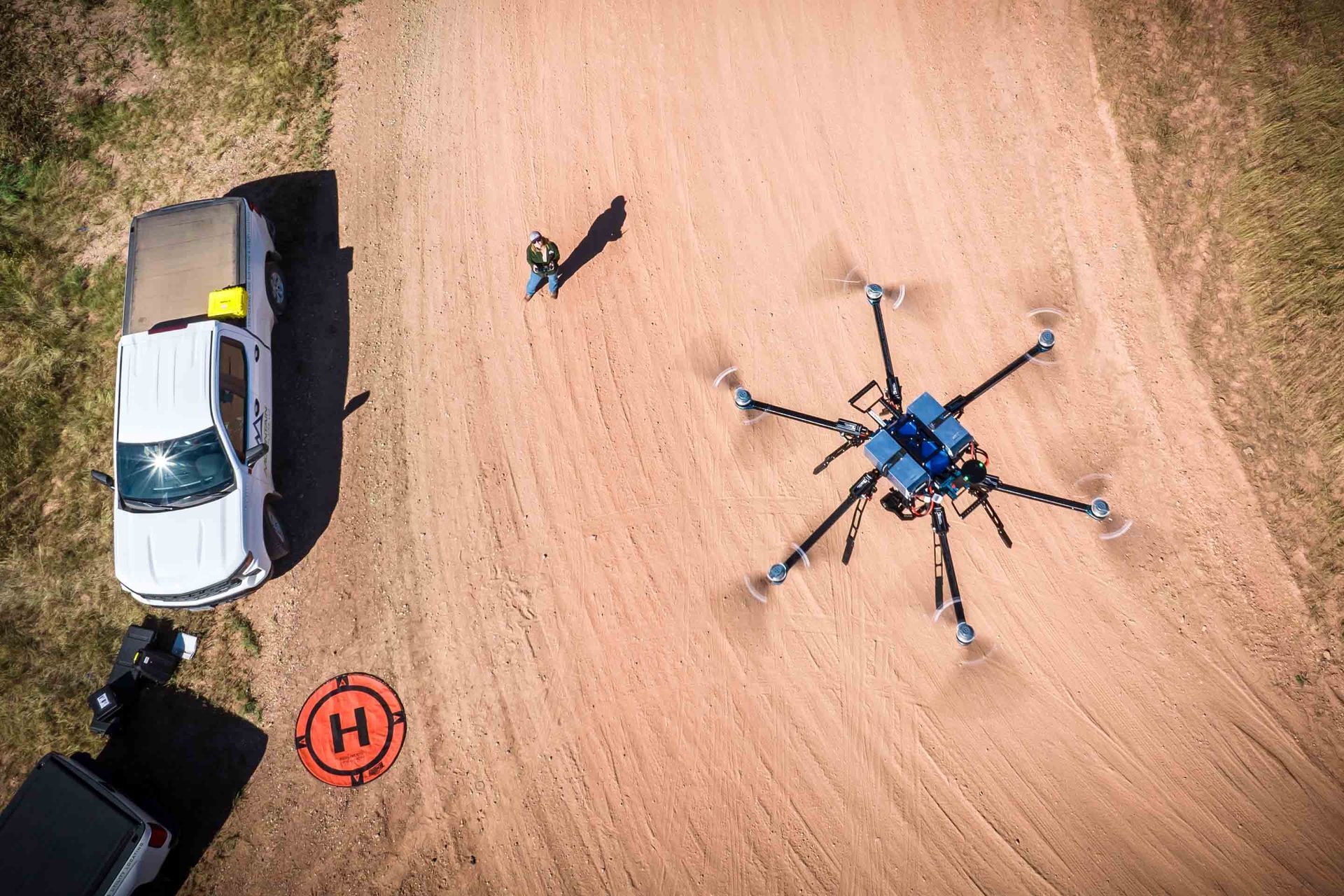 Aerial view: large drone on dirt ground with person and vehicle nearby.