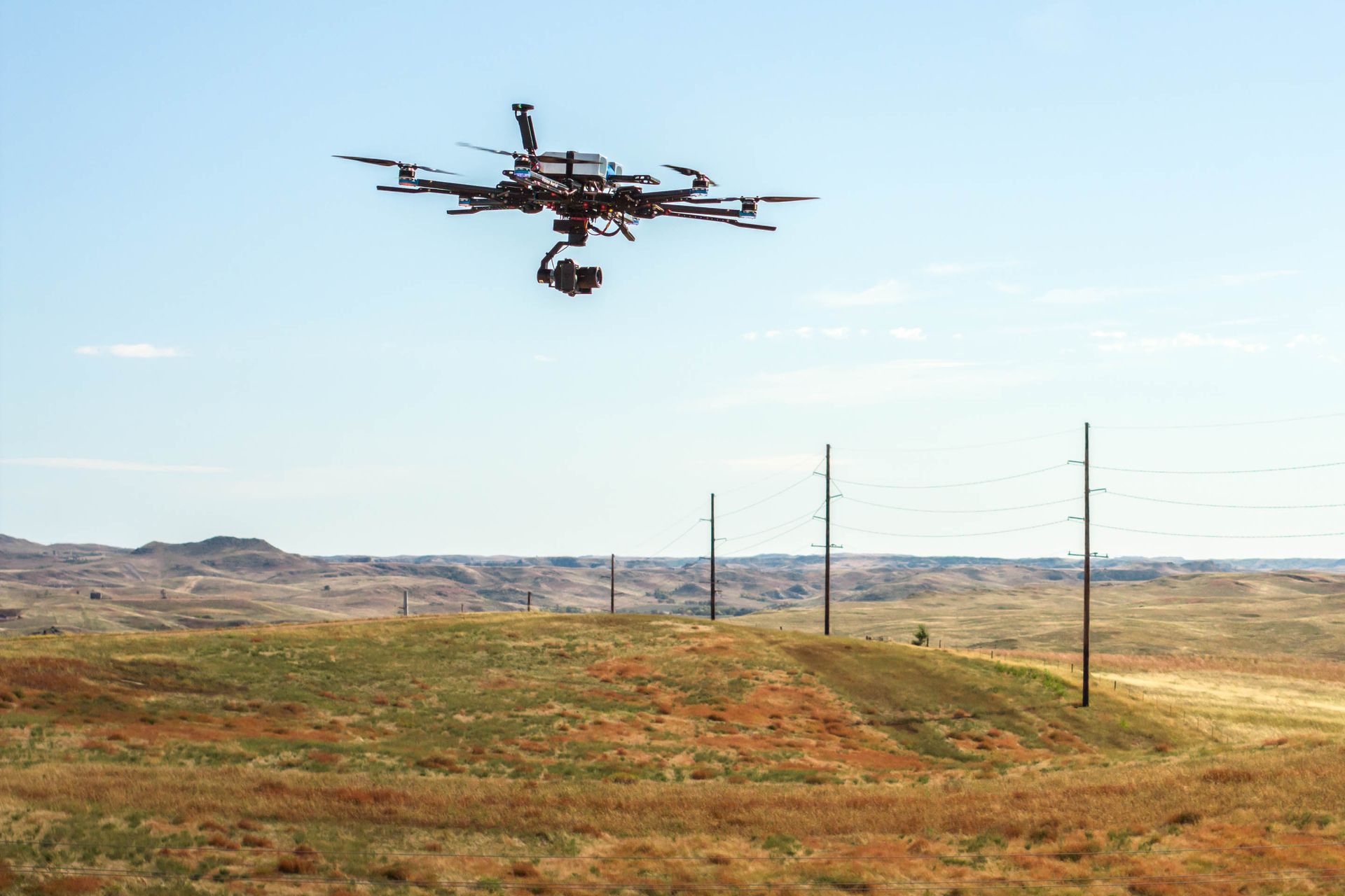 Drone flying over a grassy landscape with power lines under a blue sky.