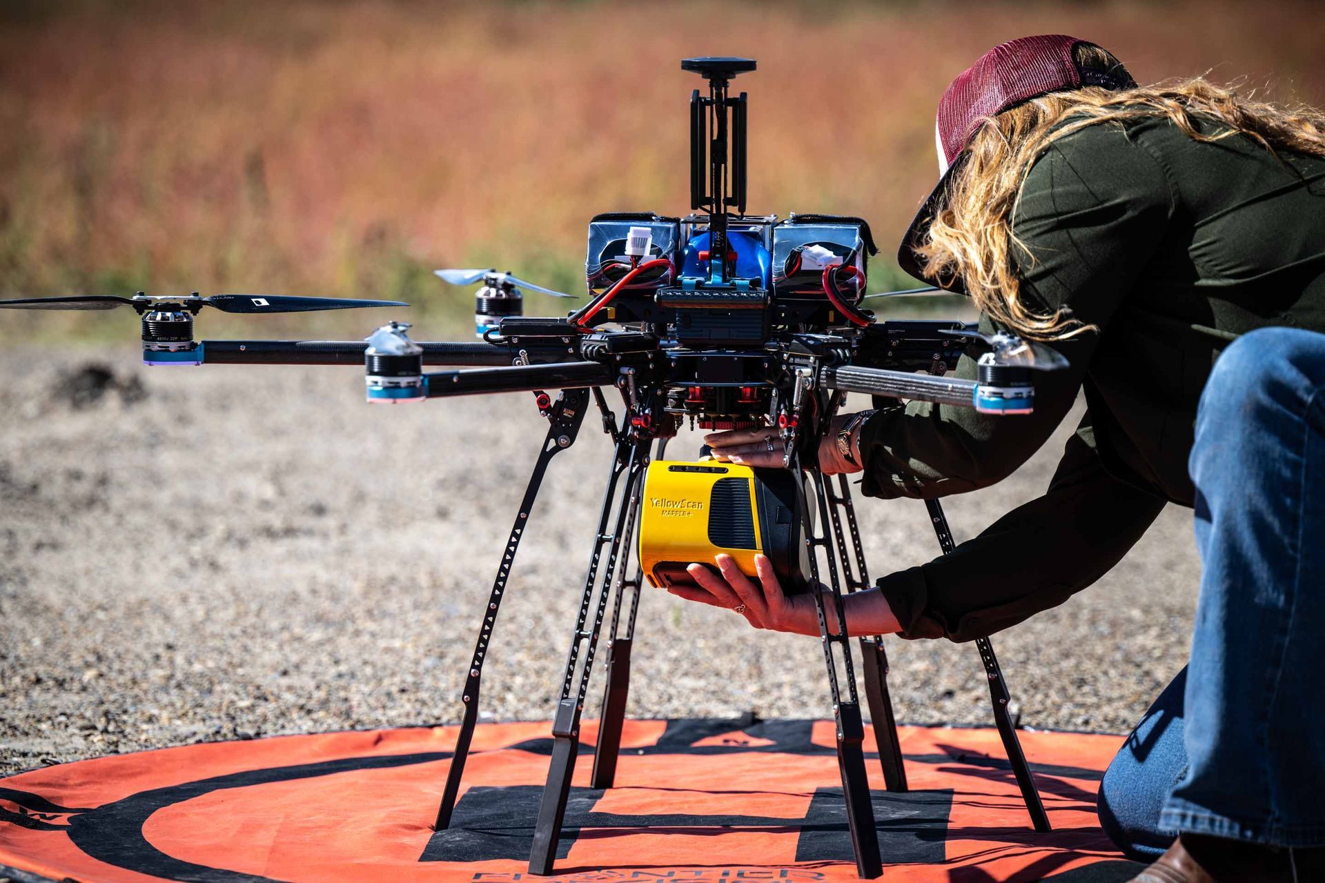 Person attaching a yellow box to a large drone on a red landing pad outdoors.