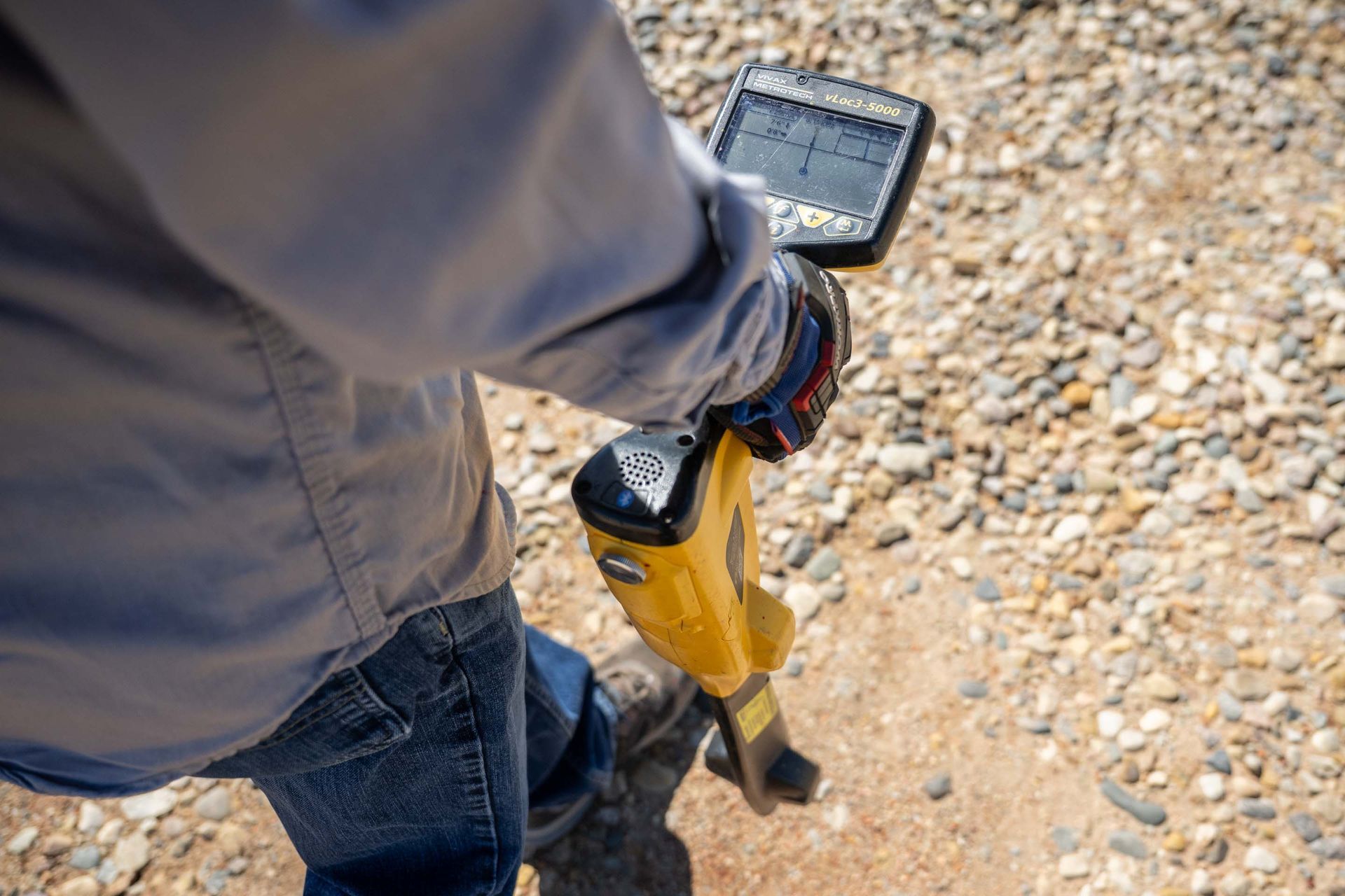Person holding a yellow utility locator device outdoors on a gravel surface.