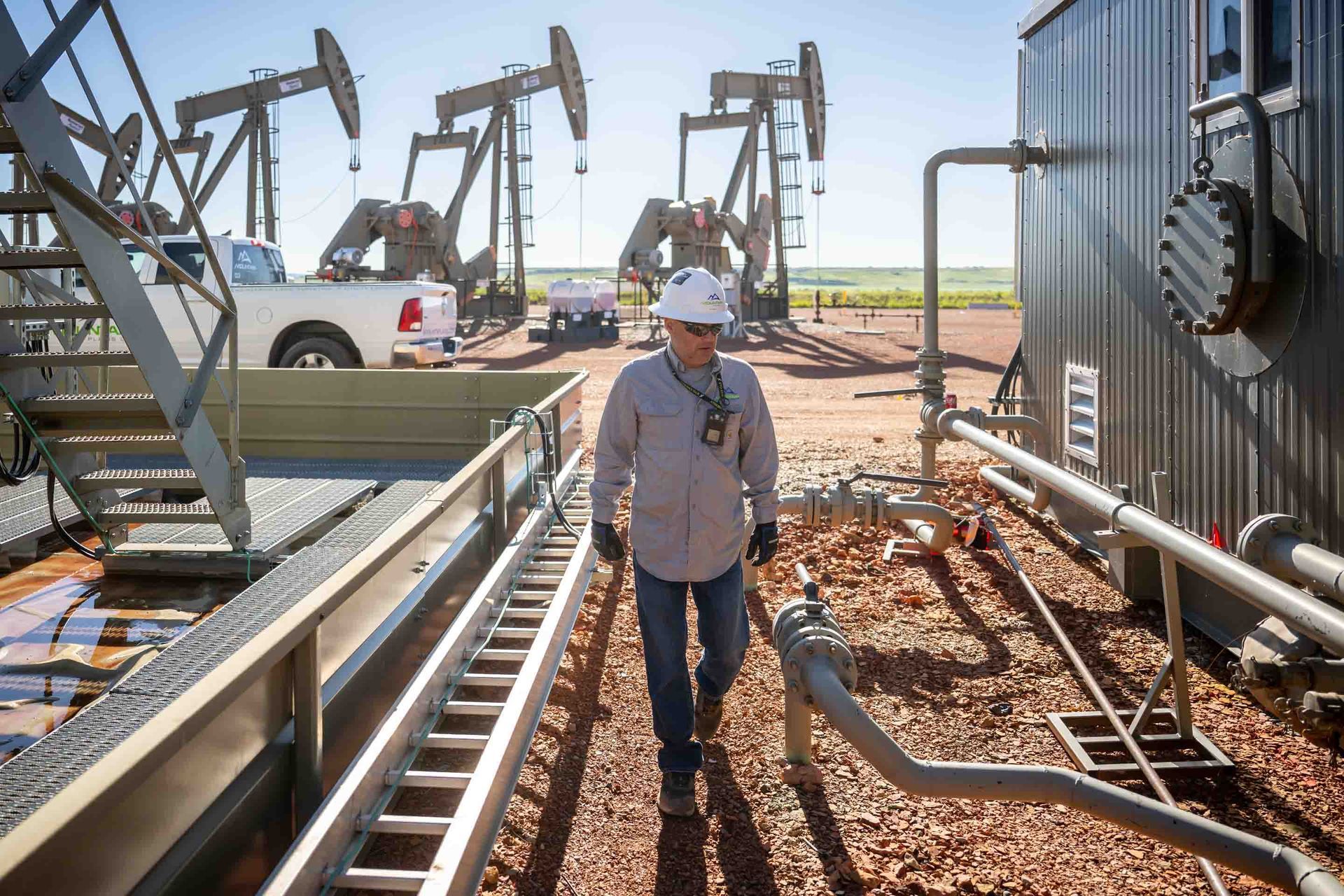 Person in a hard hat walks through an oil field, pump jacks in the background.