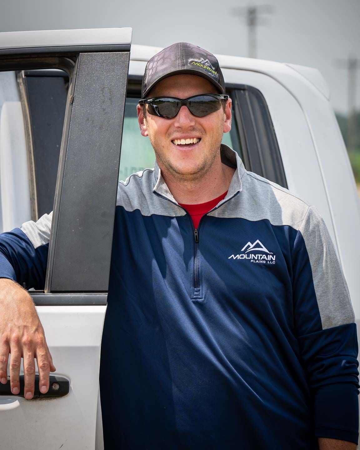 Man in sunglasses and hat smiles, leaning on a white truck. He wears a blue and gray shirt.