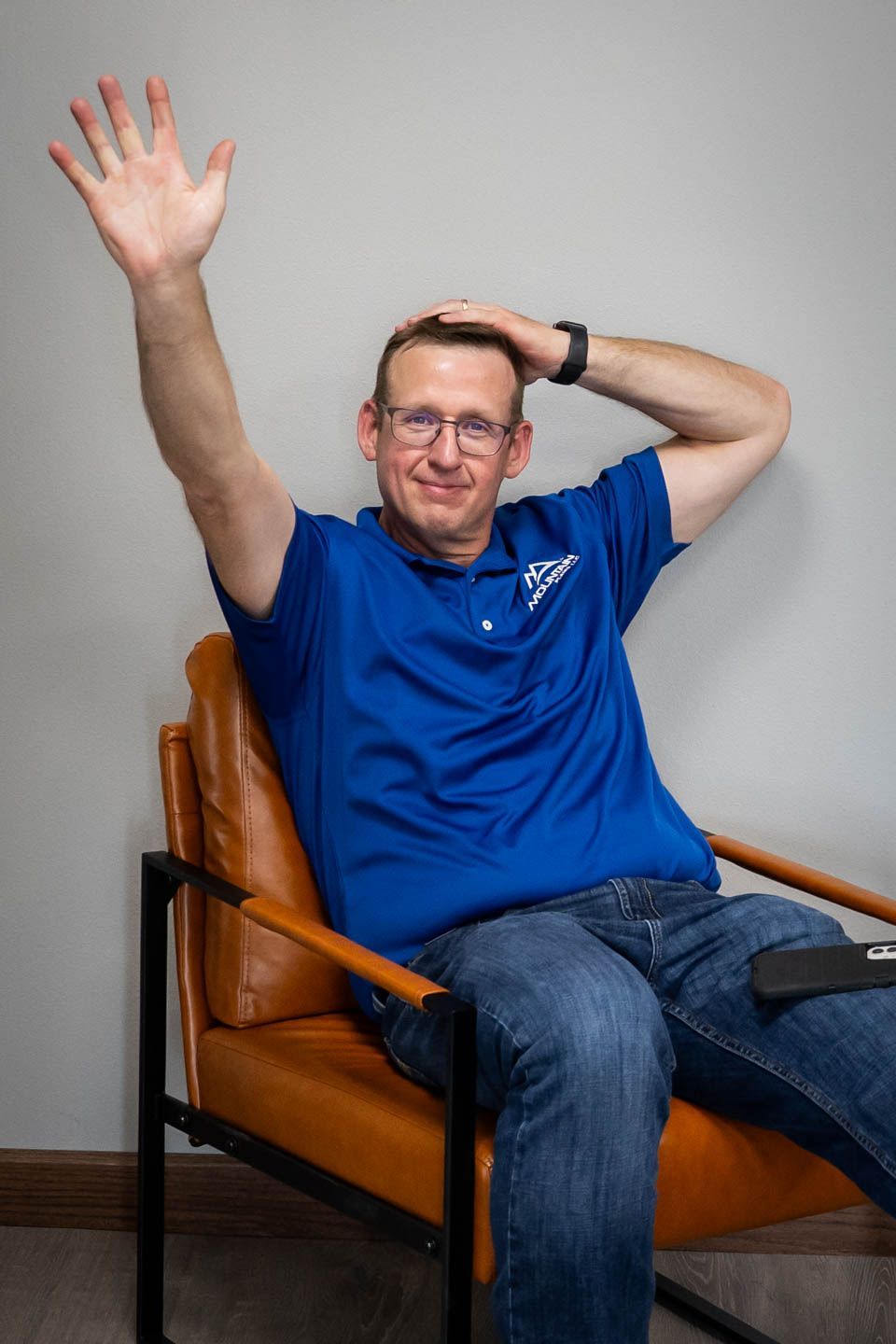Man in blue shirt waves, seated in a brown chair, gray wall in background.