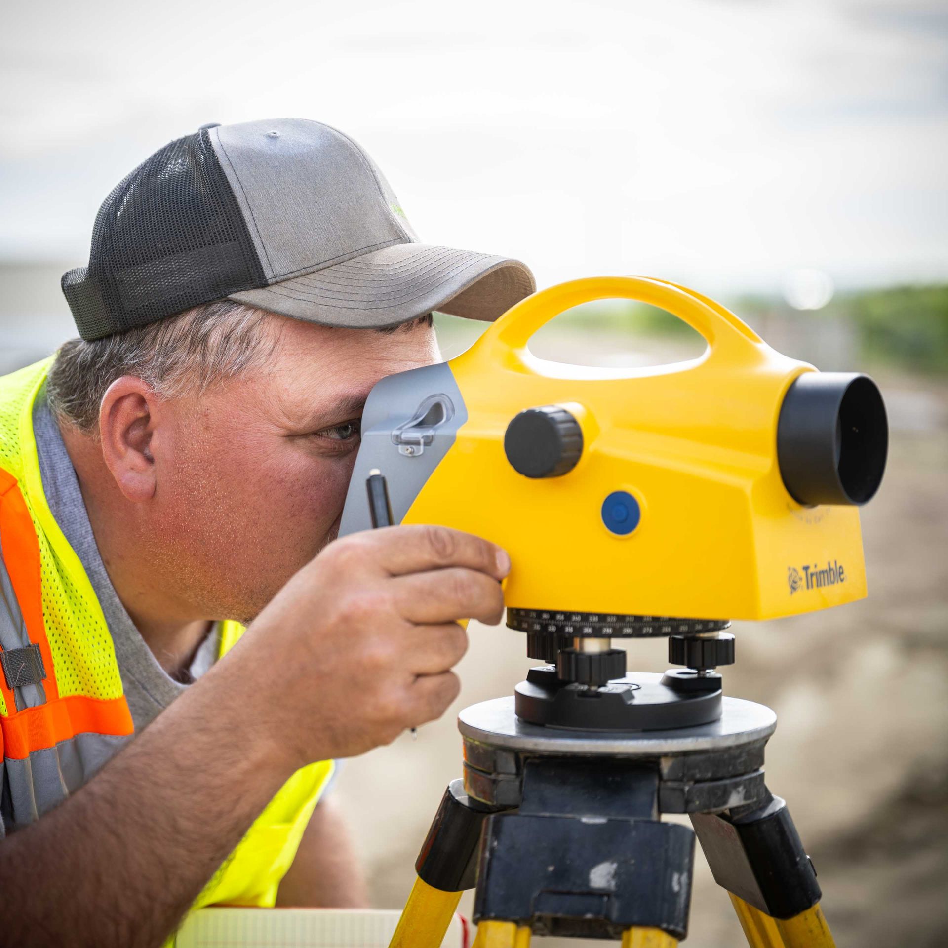 Man using yellow surveyor's level on a tripod outdoors, wearing safety vest and cap.