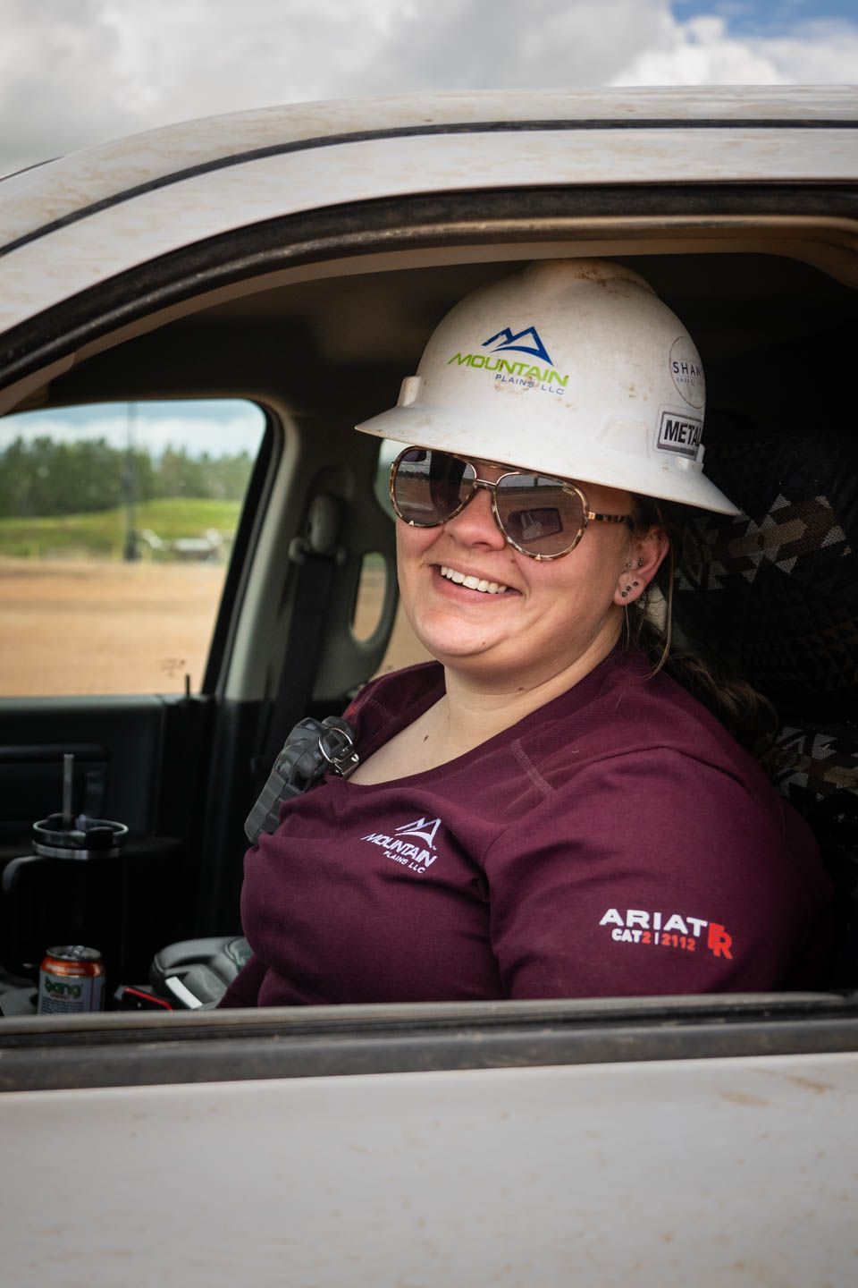 Woman in hard hat and sunglasses smiles from a vehicle, wearing a maroon shirt.