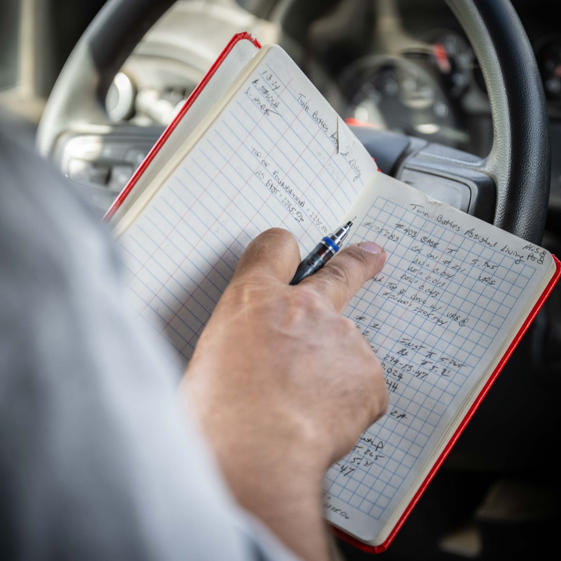 Person in a vehicle points at notes in a red notebook on the steering wheel.