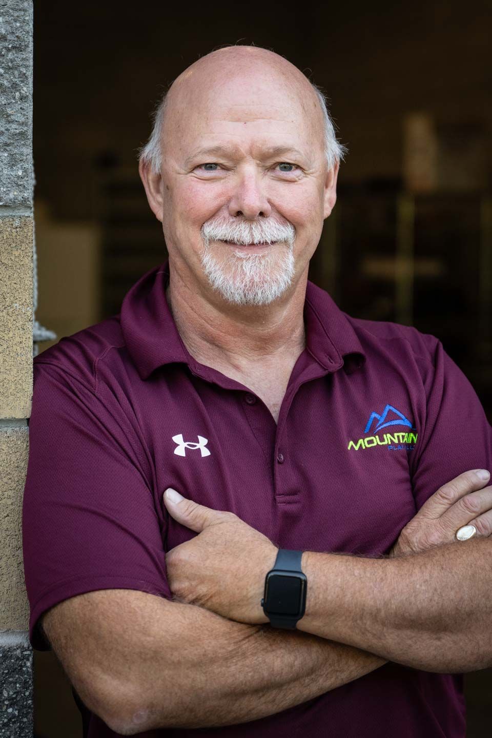 Man with a bald head and graying beard, arms crossed, wearing a maroon polo shirt with a mountain logo.
