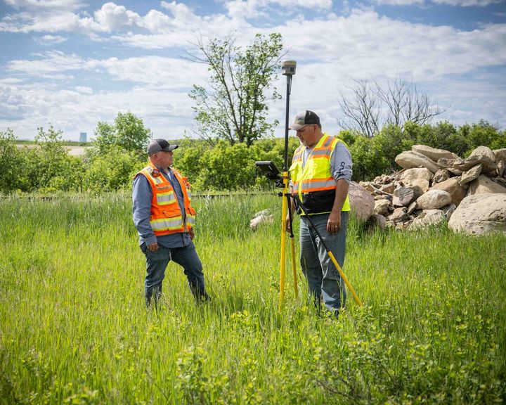 Two people in safety vests surveying a grassy field with equipment on a tripod.