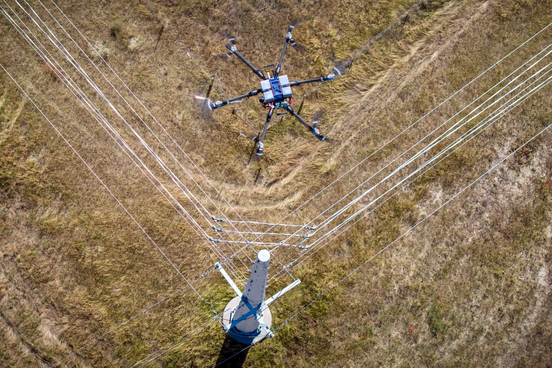 Drone inspecting power lines in a field; white cables connected to the drone and power lines are visible.