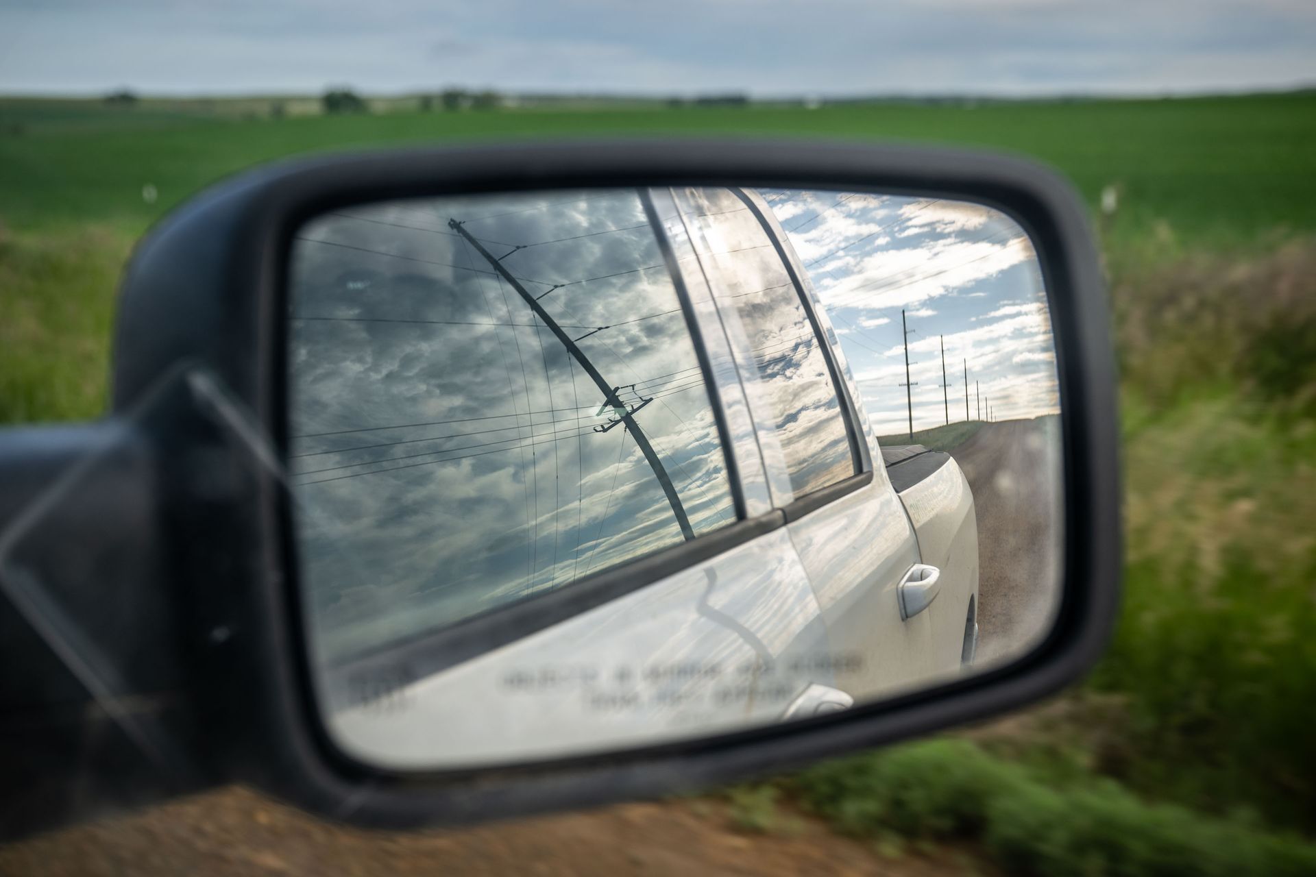 Side mirror reflecting a vehicle and cloudy sky, roadside view.
