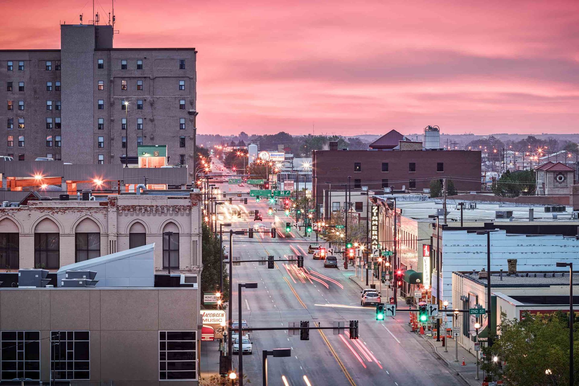 City street at dusk, with buildings and traffic lit by streetlights against a pink sky.