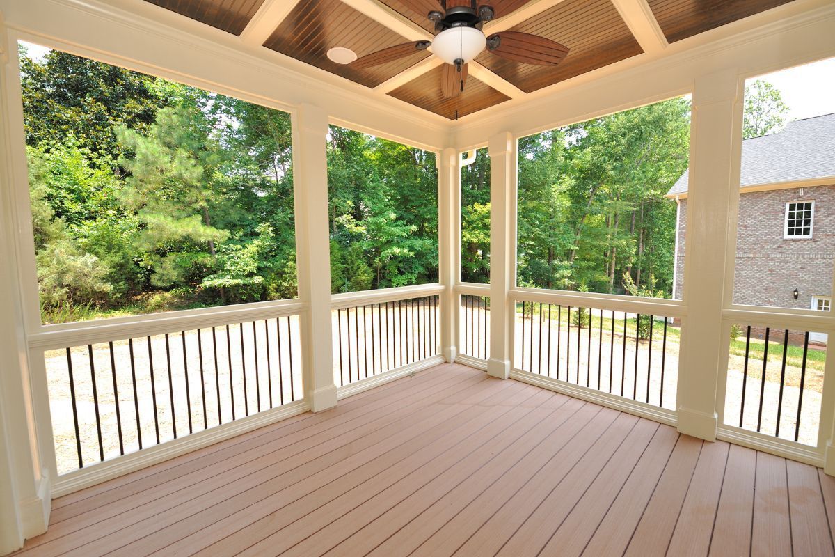 An expansive screened porch in Gilbert, AZ, featuring a wooden deck floor with a natural finish. The porch is framed by white columns and railings that support a high ceiling with a dark wood finish and a ceiling fan, blending functionality with aesthetic appeal. Large screened openings allow for an unobstructed view of the surrounding lush greenery, inviting the outdoors in while providing shade and protection from the elements. The space evokes a tranquil and inviting atmosphere, ideal for relaxation or entertaining.