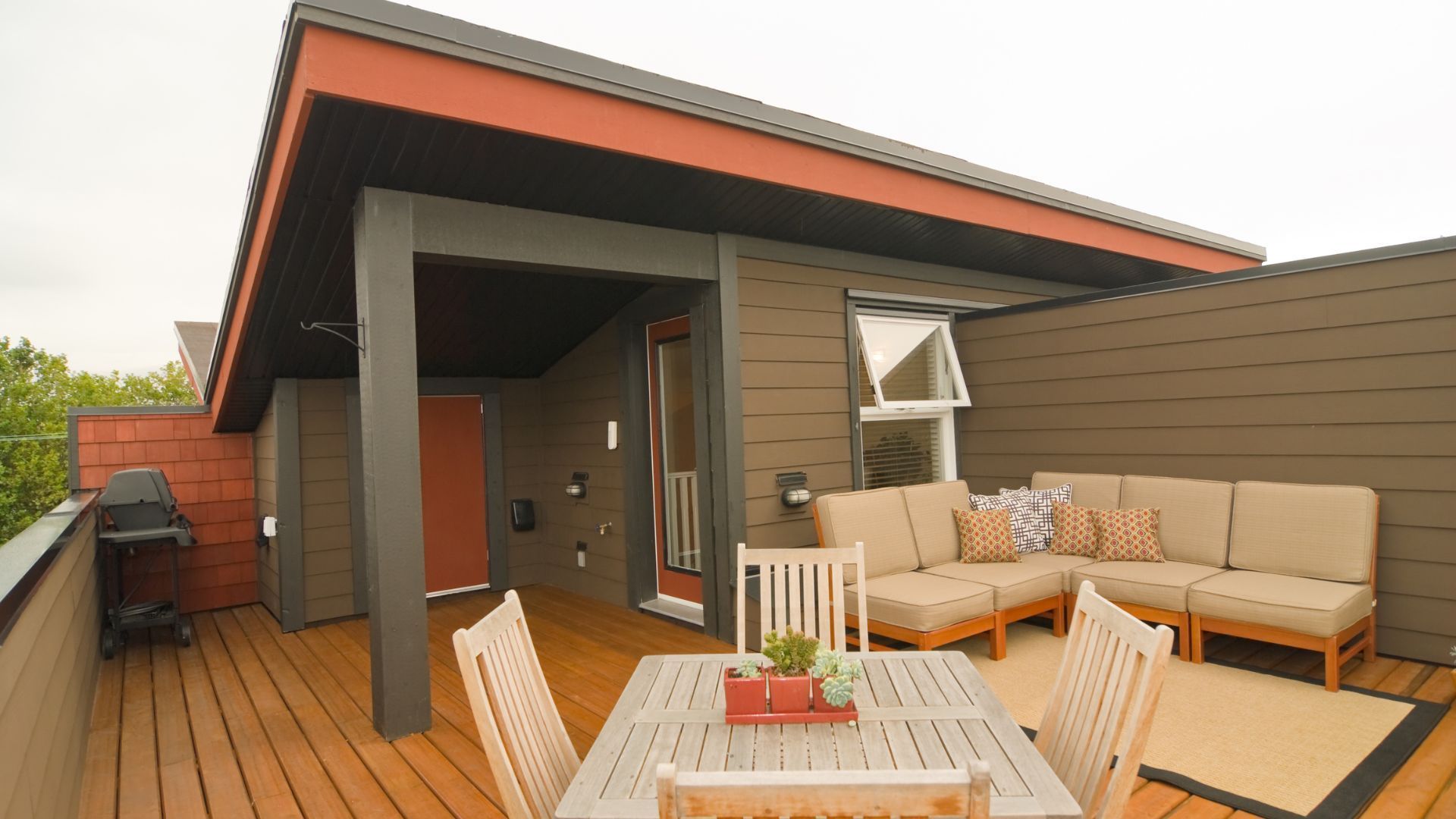 Spacious and inviting custom-designed wooden deck in Gilbert, Arizona, featuring comfortable outdoor furniture set with plush beige cushions and colorful throw pillows. A sturdy wooden table with a set of succulents centers the space, while a barbecue grill stands ready for outdoor cooking. The deck's warm wooden tones contrast beautifully with the home's modern grey siding and vibrant red-orange trim, embodying Gilbert’s sunny, desert aesthetic. The covered patio area promises an ideal spot for enjoying the Arizona climate in a stylish, private backyard setting.