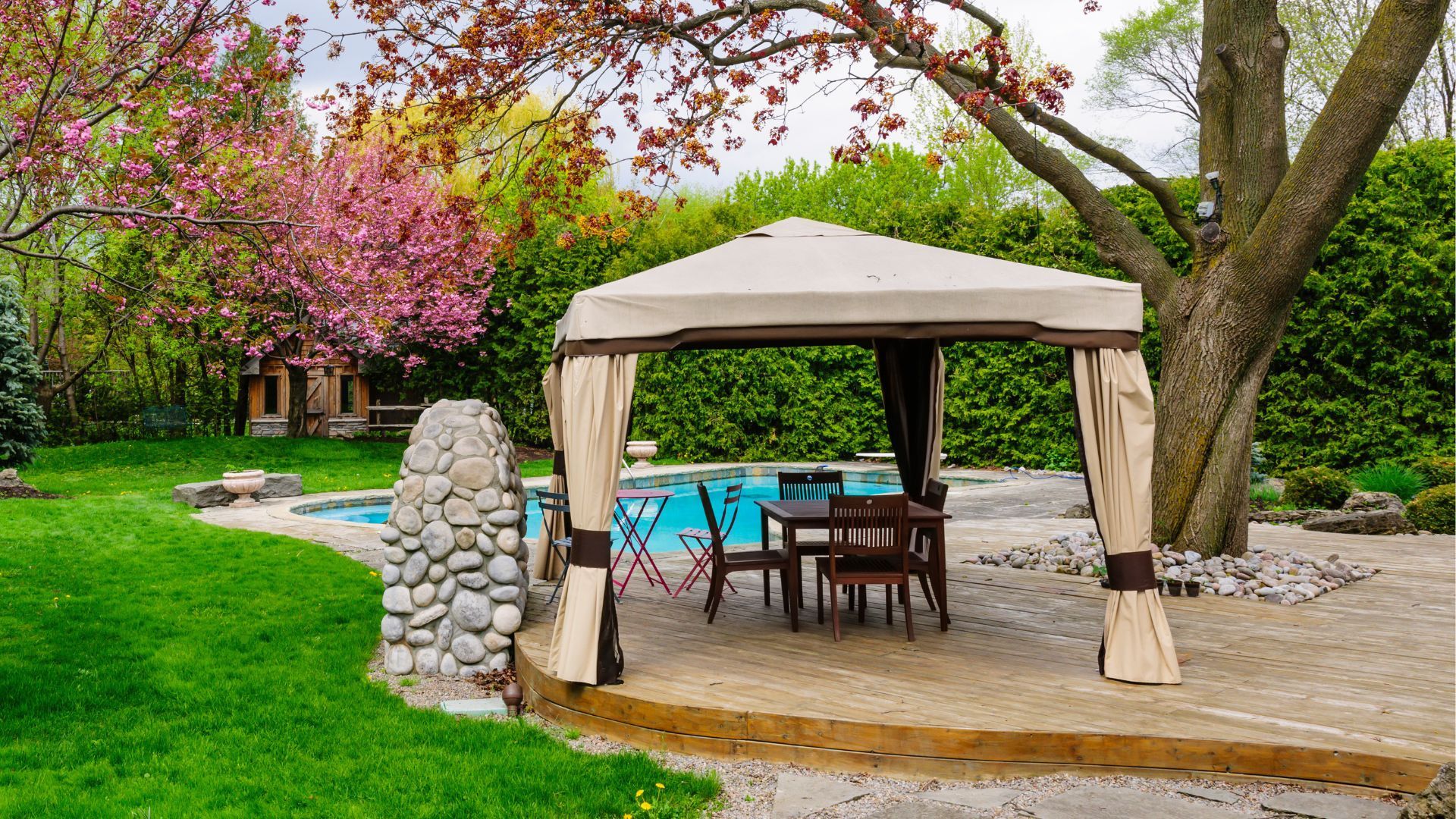 Wooden deck with gazebo, pool, and blooming cherry tree in a lush garden.