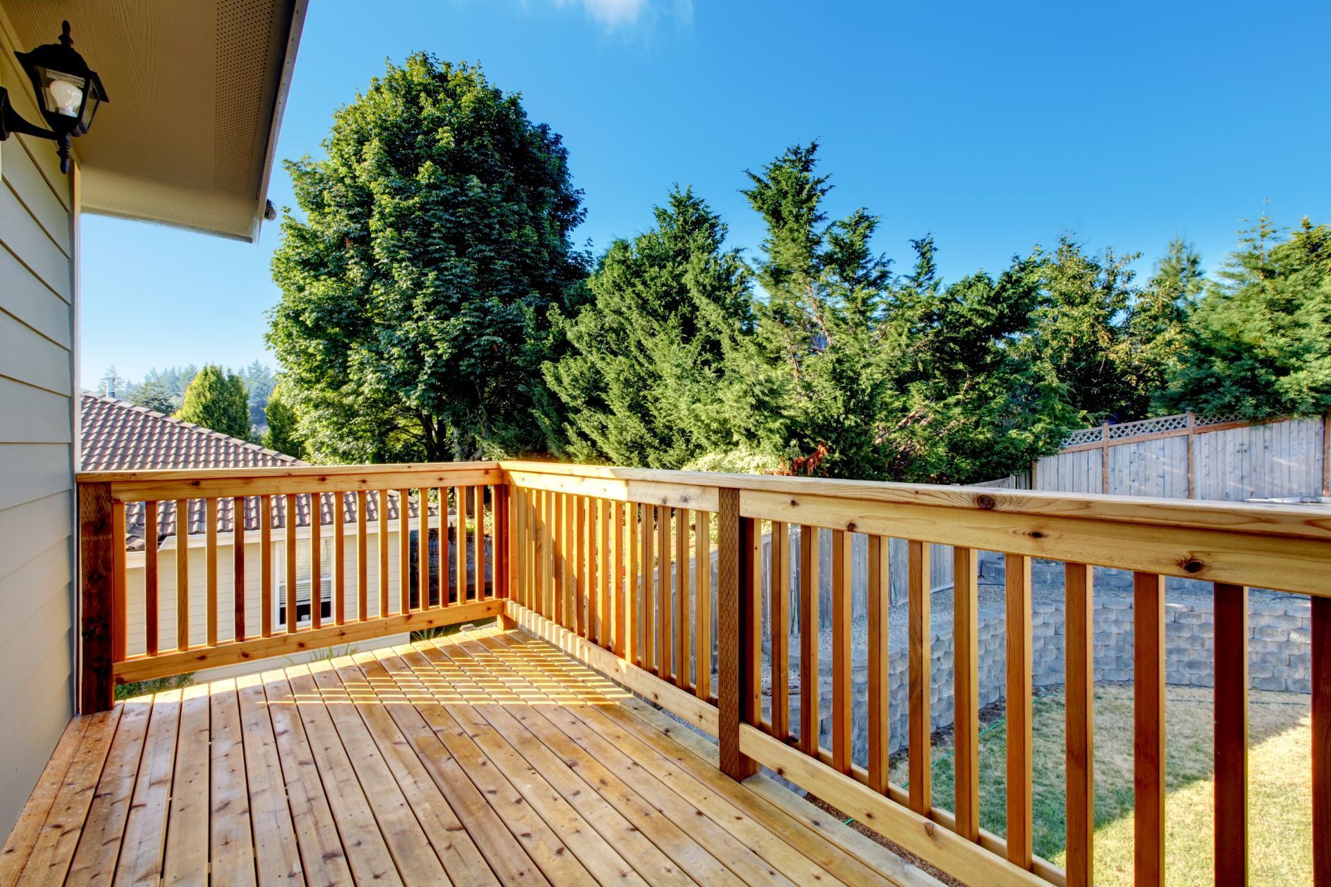 A wooden deck bathed in sunlight features a natural finish and is enclosed with a matching wooden railing in Gilbert, AZ. The deck overlooks a backyard bordered by dense, tall trees and a classic wooden fence, creating a private and tranquil outdoor space. The shadow pattern of the railing decorates the deck floor, adding to the inviting ambiance of this suburban retreat.