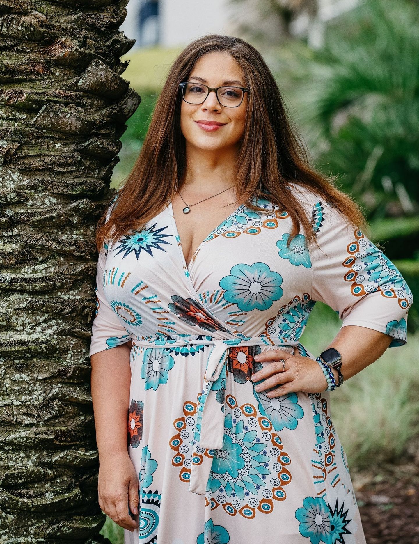 A woman in a floral dress and glasses is standing next to a palm tree.