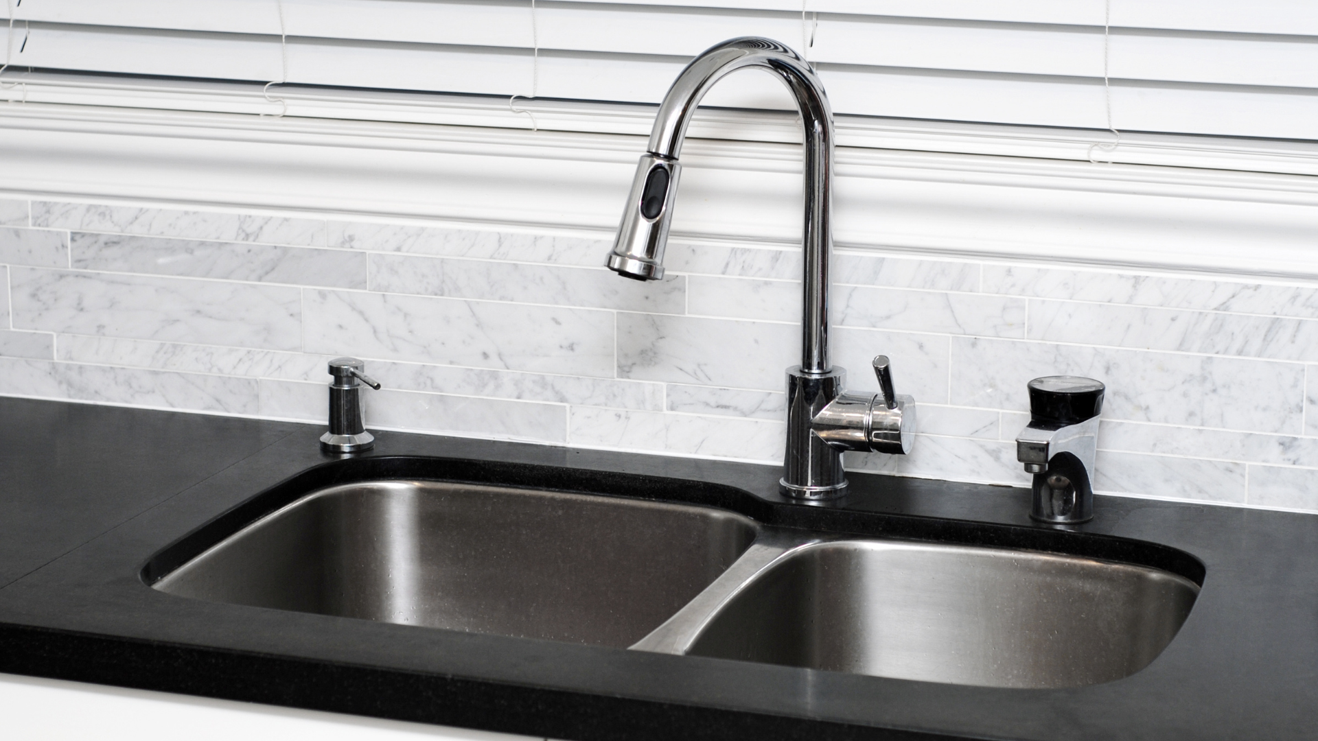 Stainless steel kitchen sink with faucet, soap dispenser, and window blinds.