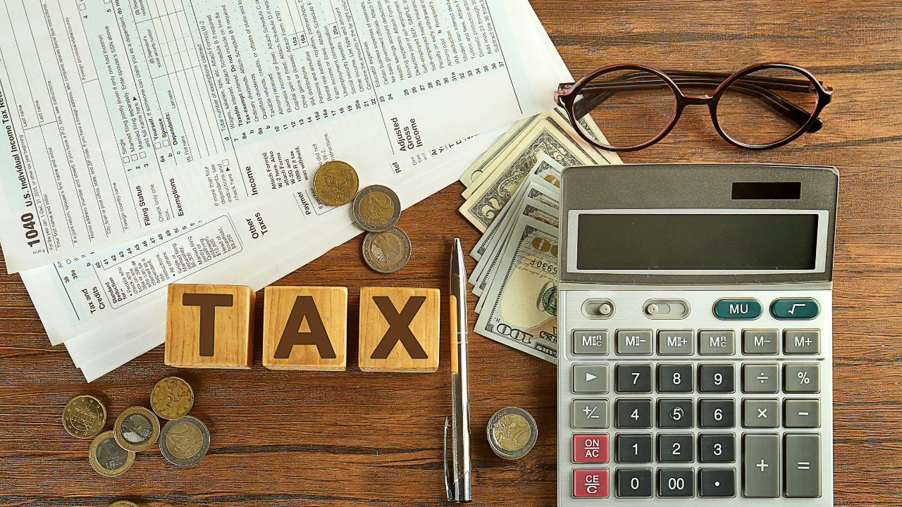 Tax documents, wooden TAX blocks, coins, calculator, pen, and eyeglasses on a desk.