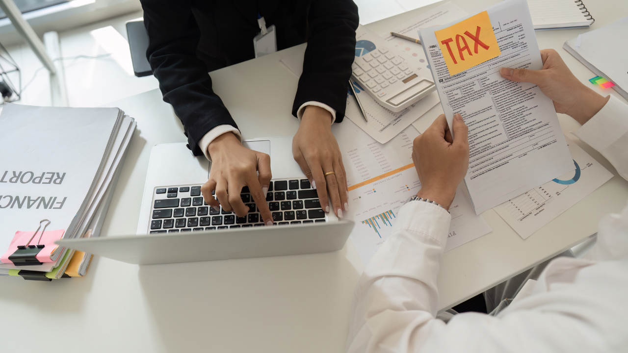 People working at a desk with a laptop and papers, reviewing a tax form labeled “TAX.”