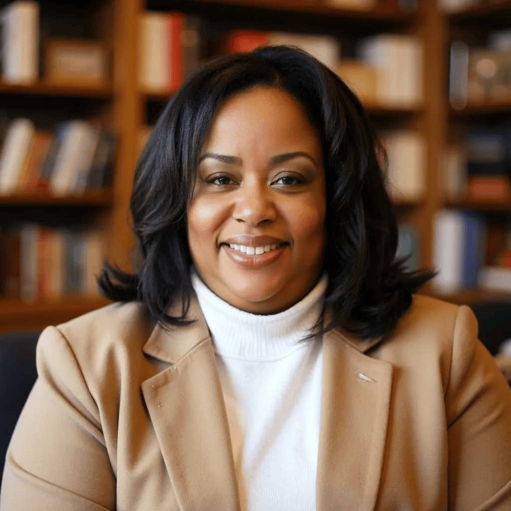 Smiling person in a tan blazer and white turtleneck seated before bookshelves