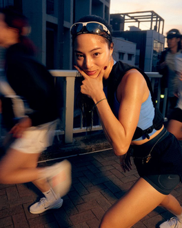 Woman in workout clothes poses, pointing at her cheek, with runners blurred in the background.
