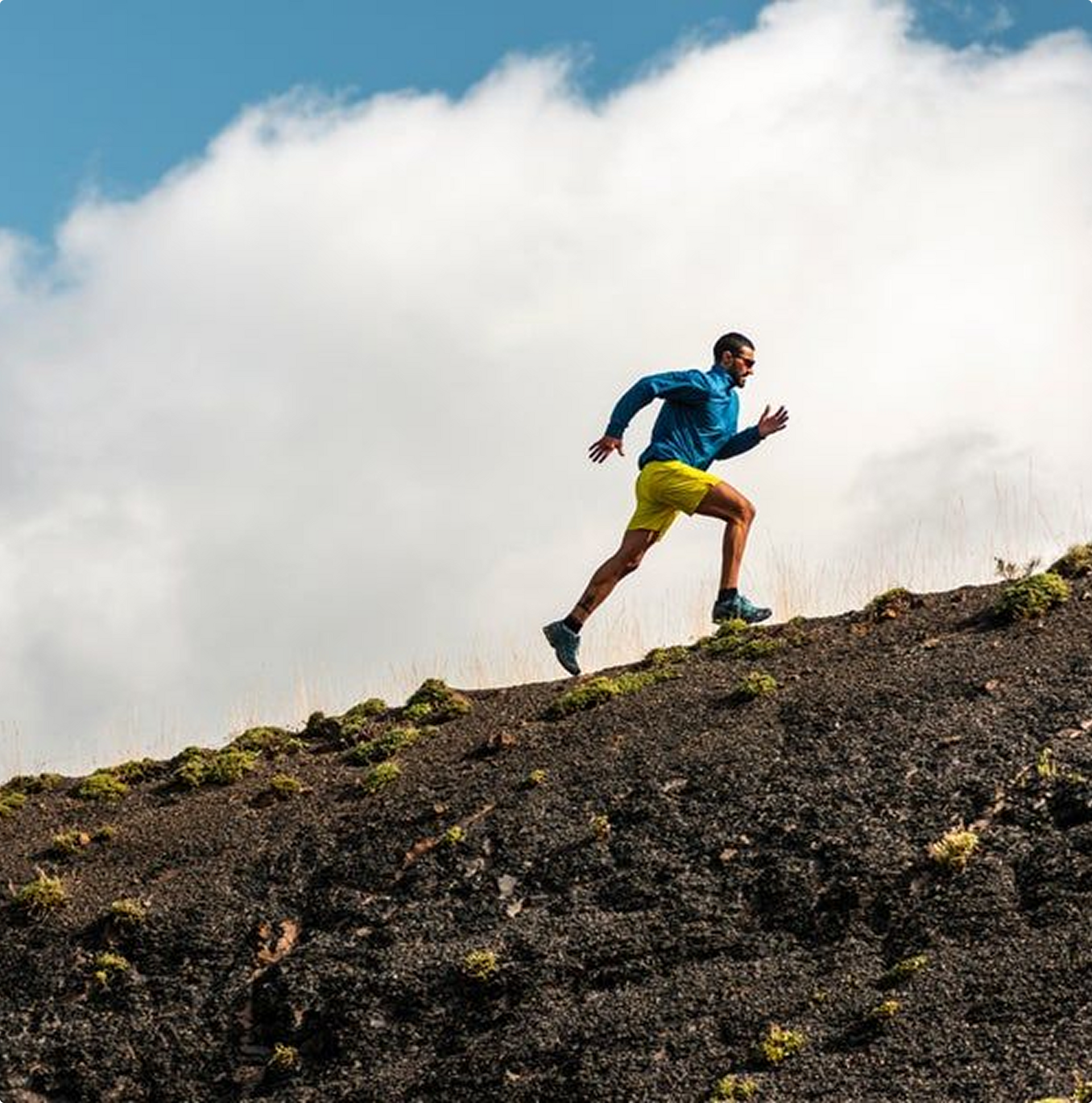 Man running uphill on a dark, rocky terrain in blue jacket, yellow shorts, with a cloudy sky.