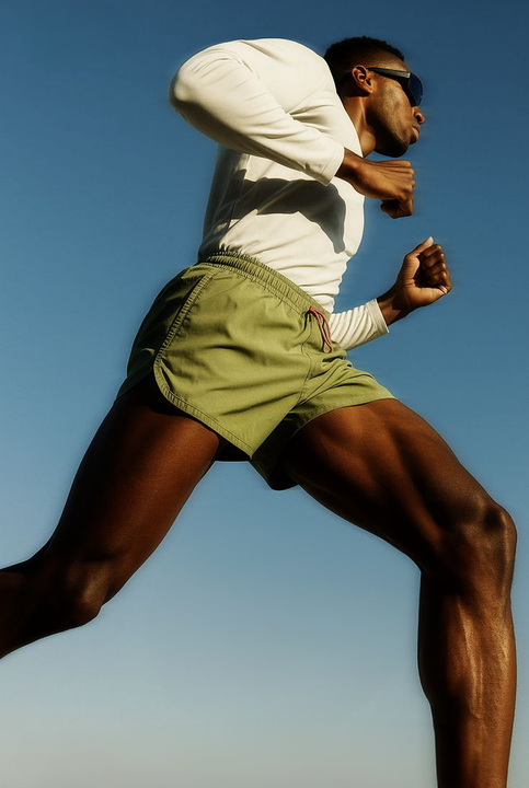 Runner, black man in green shorts, white shirt, sunglasses, mid-stride, legs extended, blue sky background.